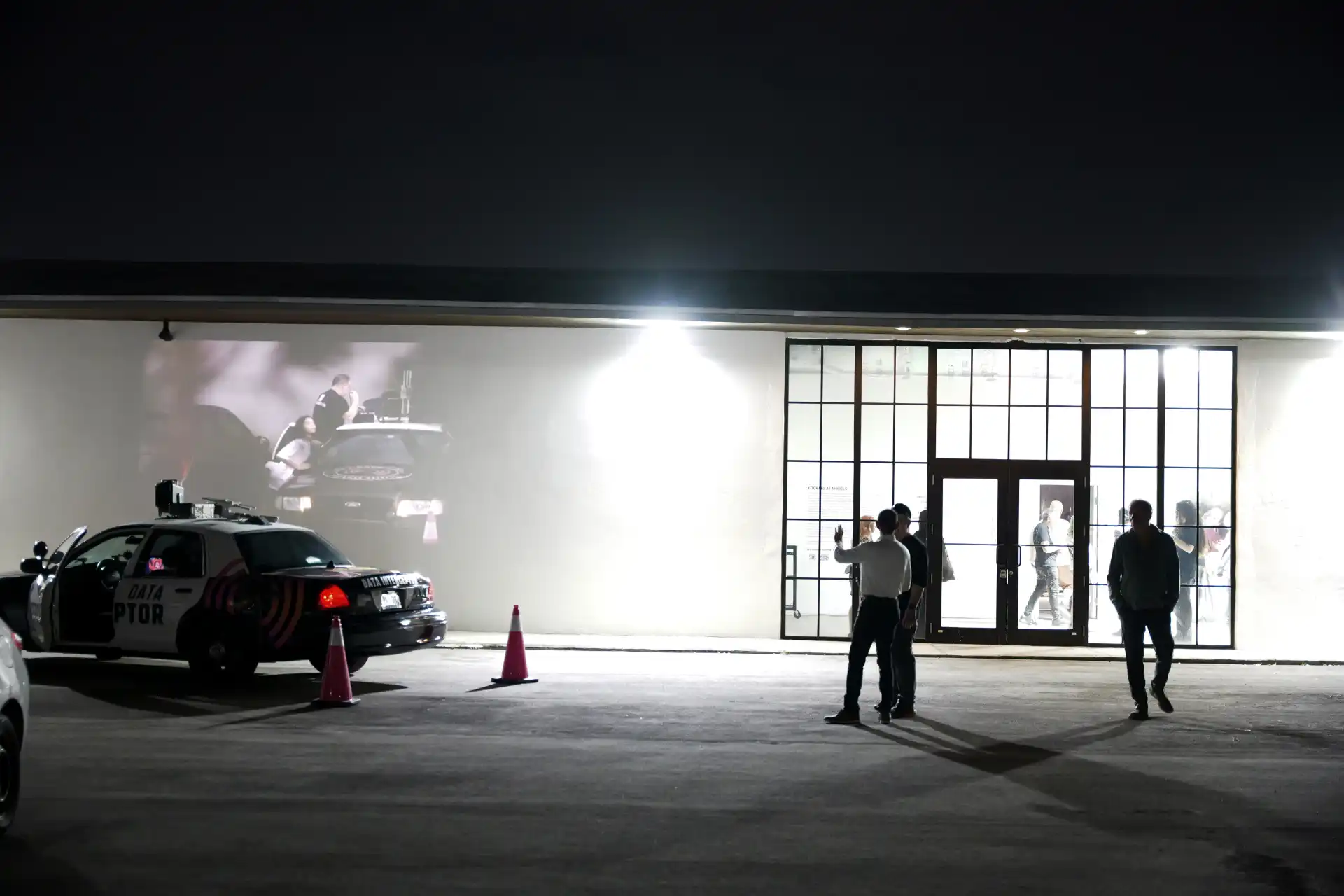 Night scene with a car projecting a video on a building wall and three people standing nearby.