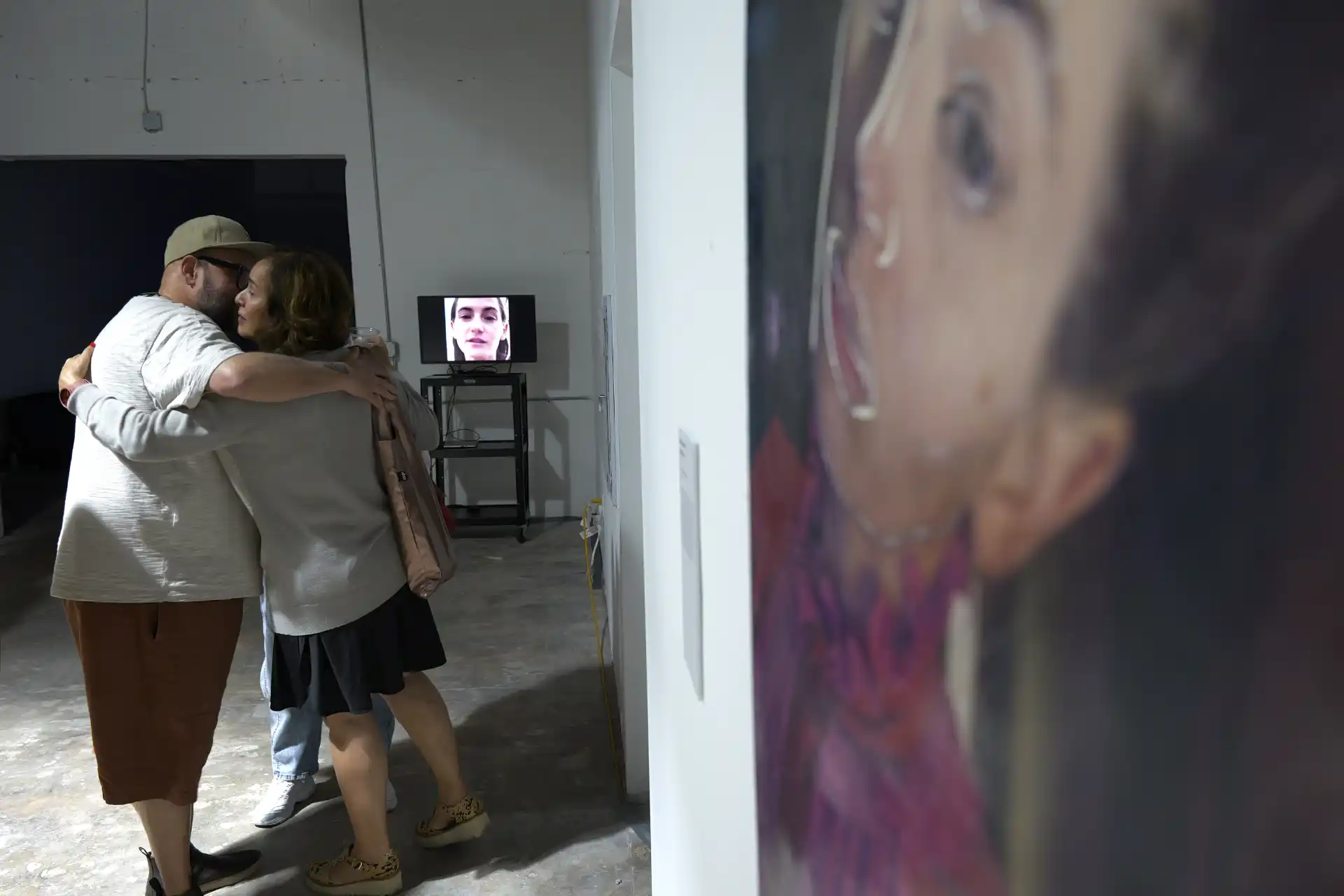 Two people embracing in a gallery with a blurred portrait painting and a video screen showing a face.