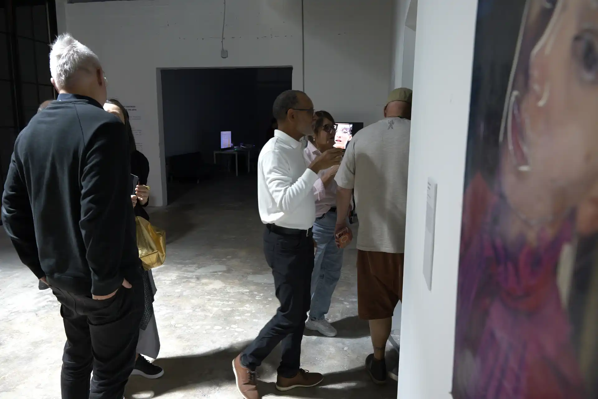 People talking and viewing art in a dimly lit gallery with a close-up portrait on the right wall.