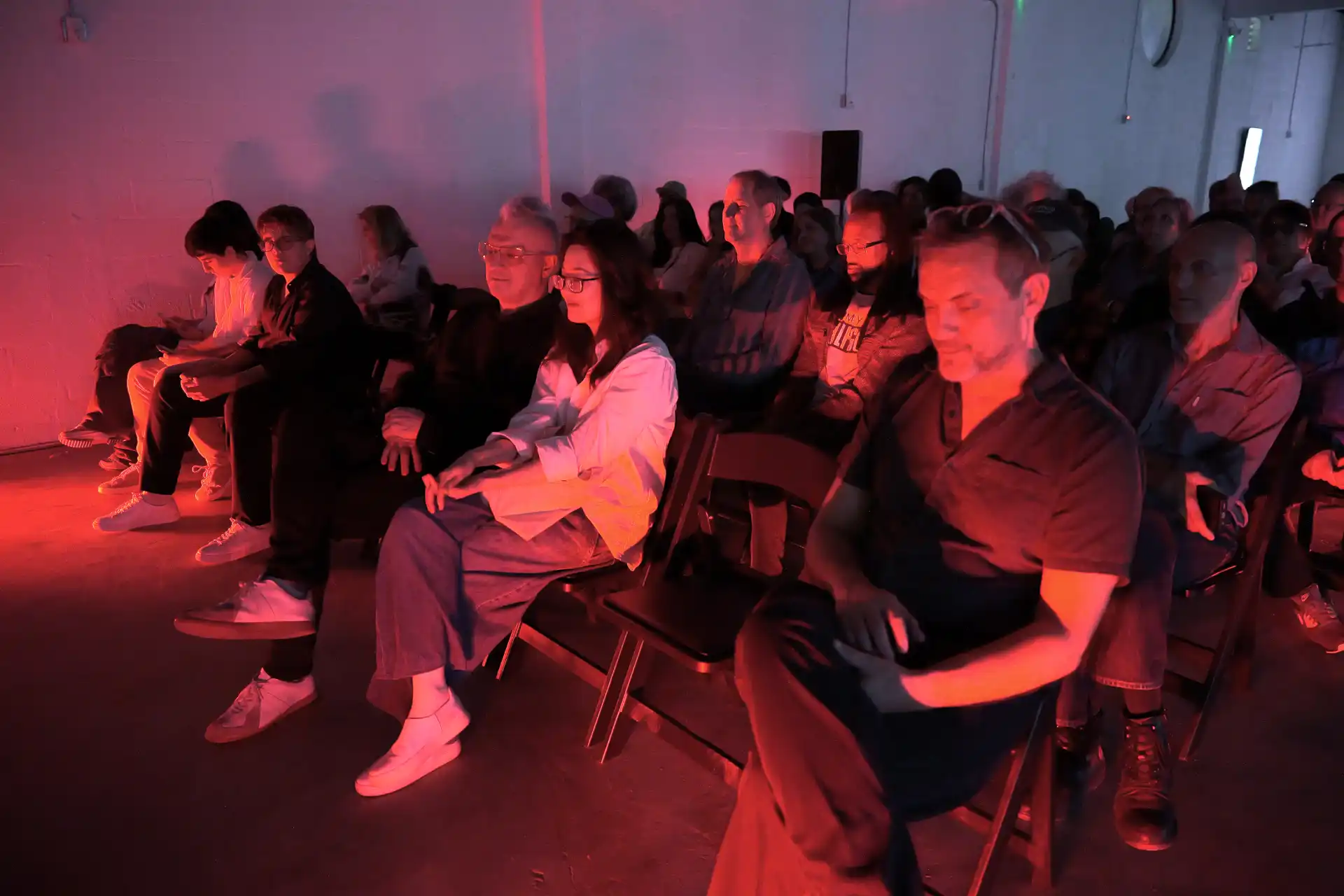 Audience seated in rows facing forward, bathed in red lighting, in a dim indoor event space.