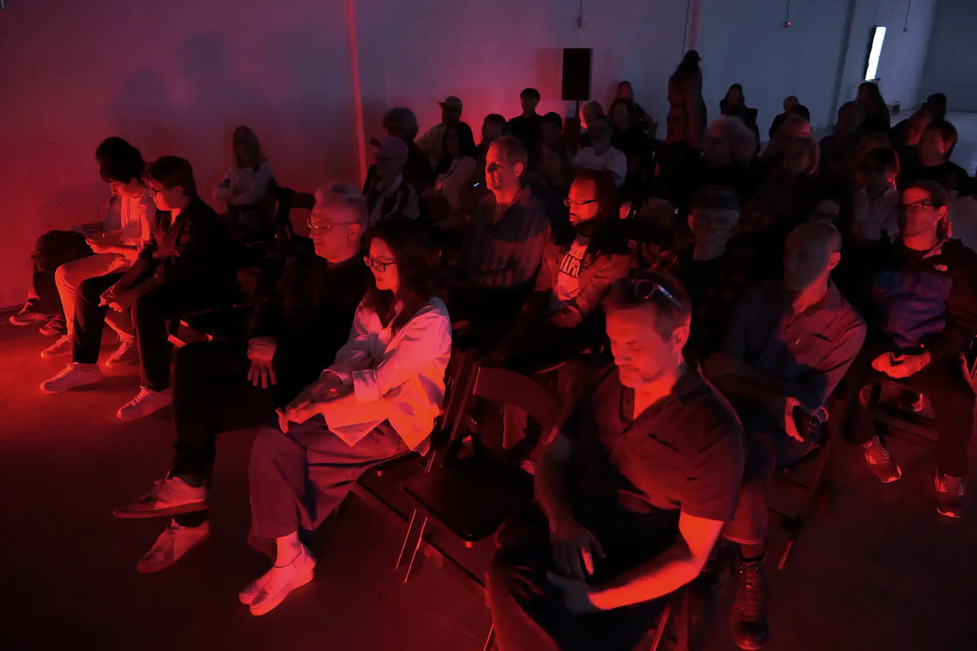 Audience seated in a dark room lit by red light, watching a performance or presentation.