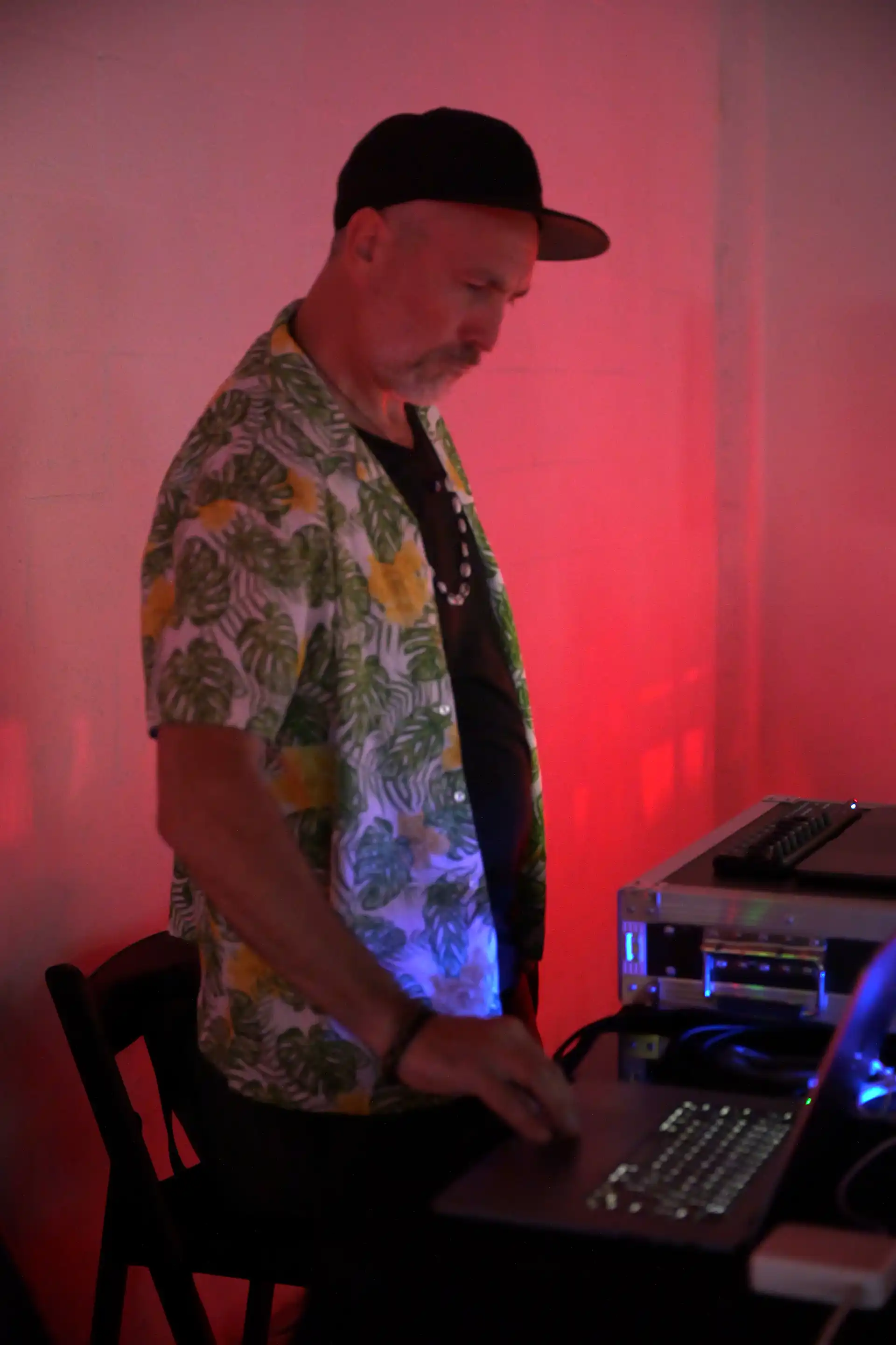 Man in floral shirt and black cap working on a laptop in a dimly lit room with red lighting.