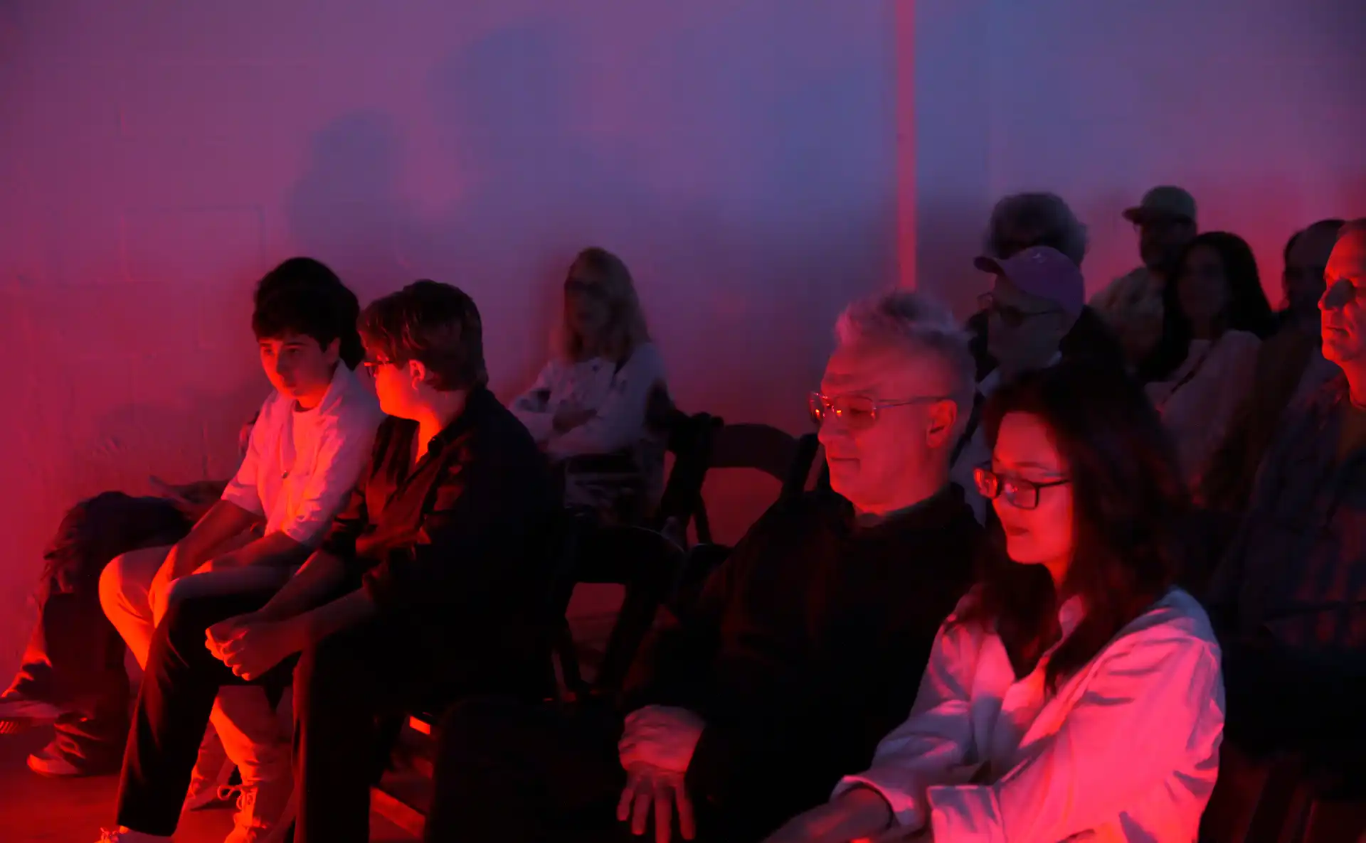 Audience sitting in dim red and purple lighting, attentively watching an event or performance.