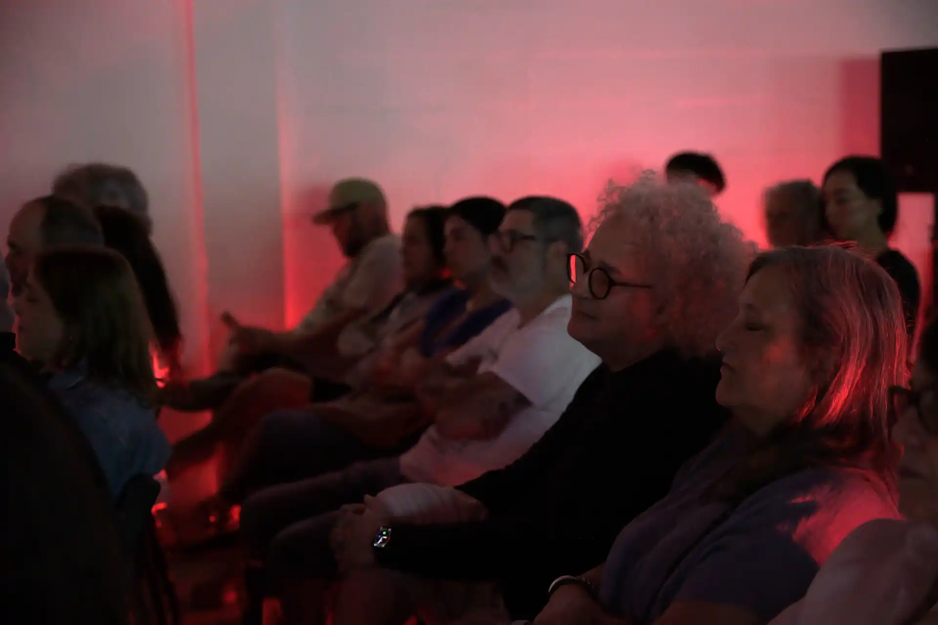 Audience seated in a dim room with red lighting, some people have eyes closed and appear relaxed.
