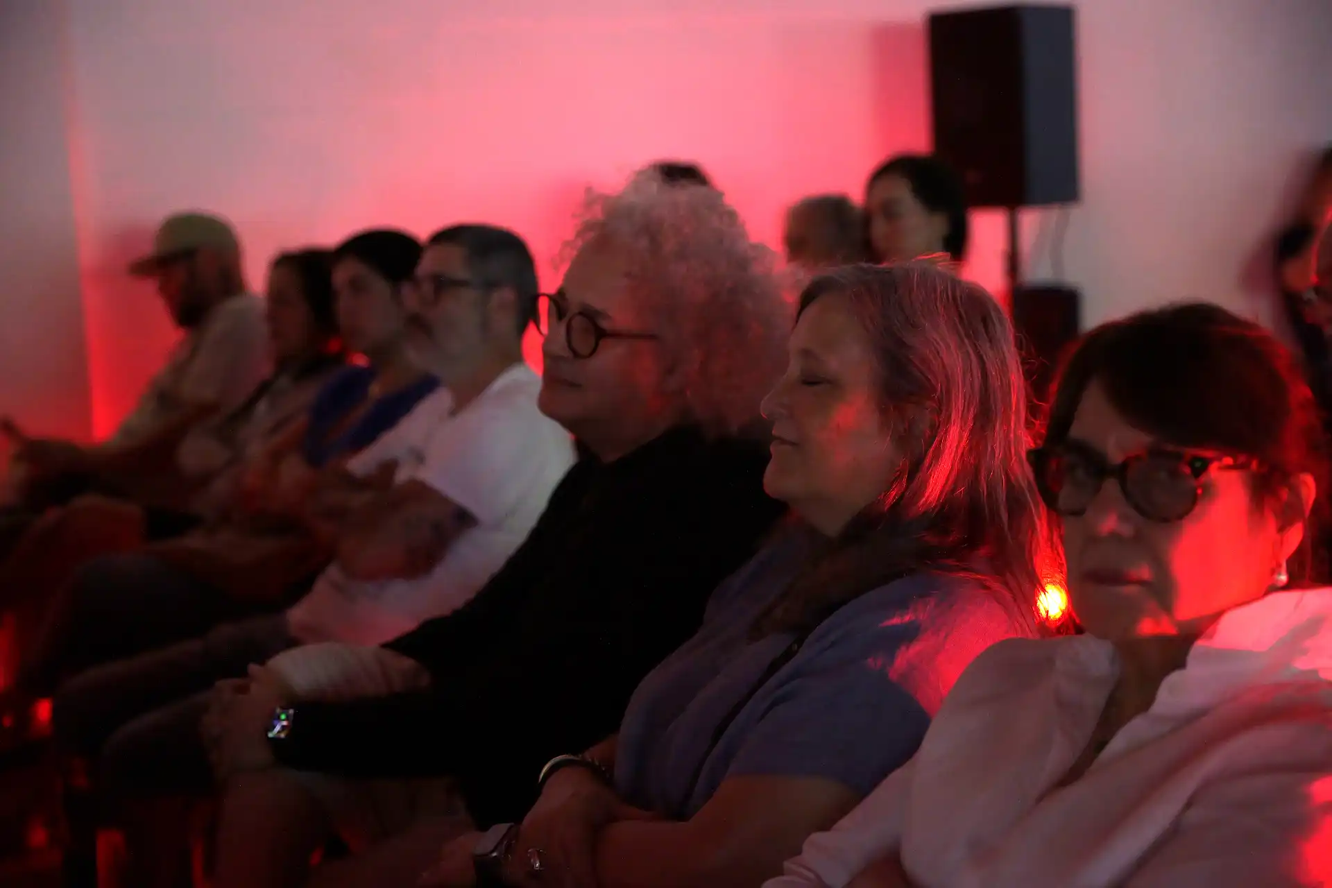 People sitting in a row indoors with red lighting, some with eyes closed, appearing relaxed or attentive.