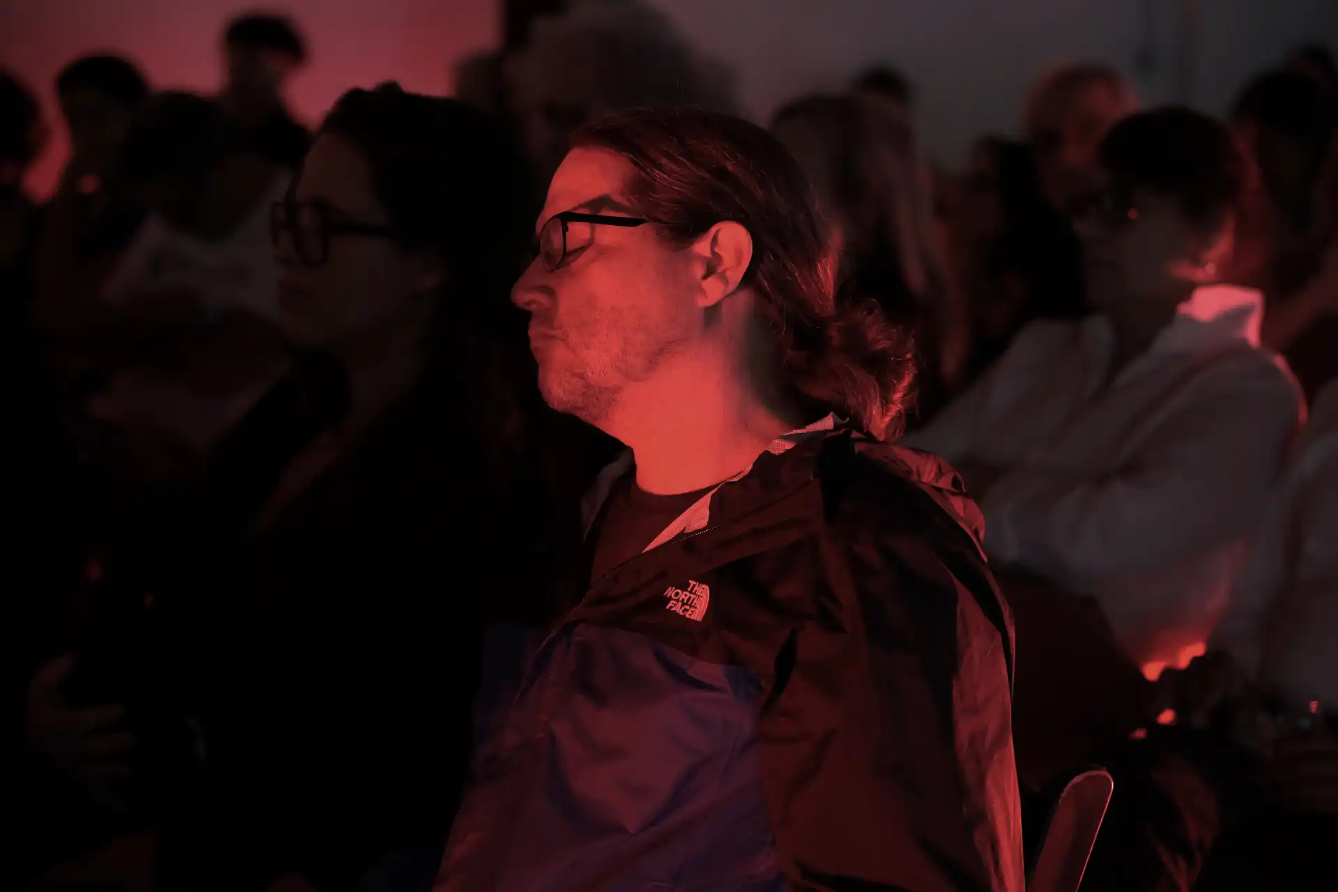 Man with glasses and ponytail sits in dark room with red lighting among others in audience.