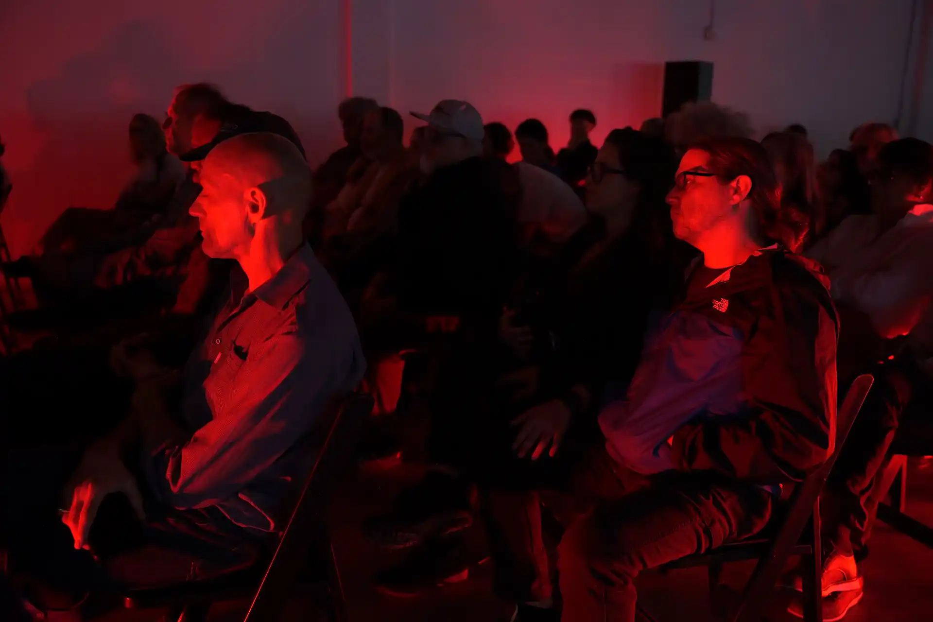 Audience seated in a dimly lit room with red lighting, attentively watching a presentation or event.