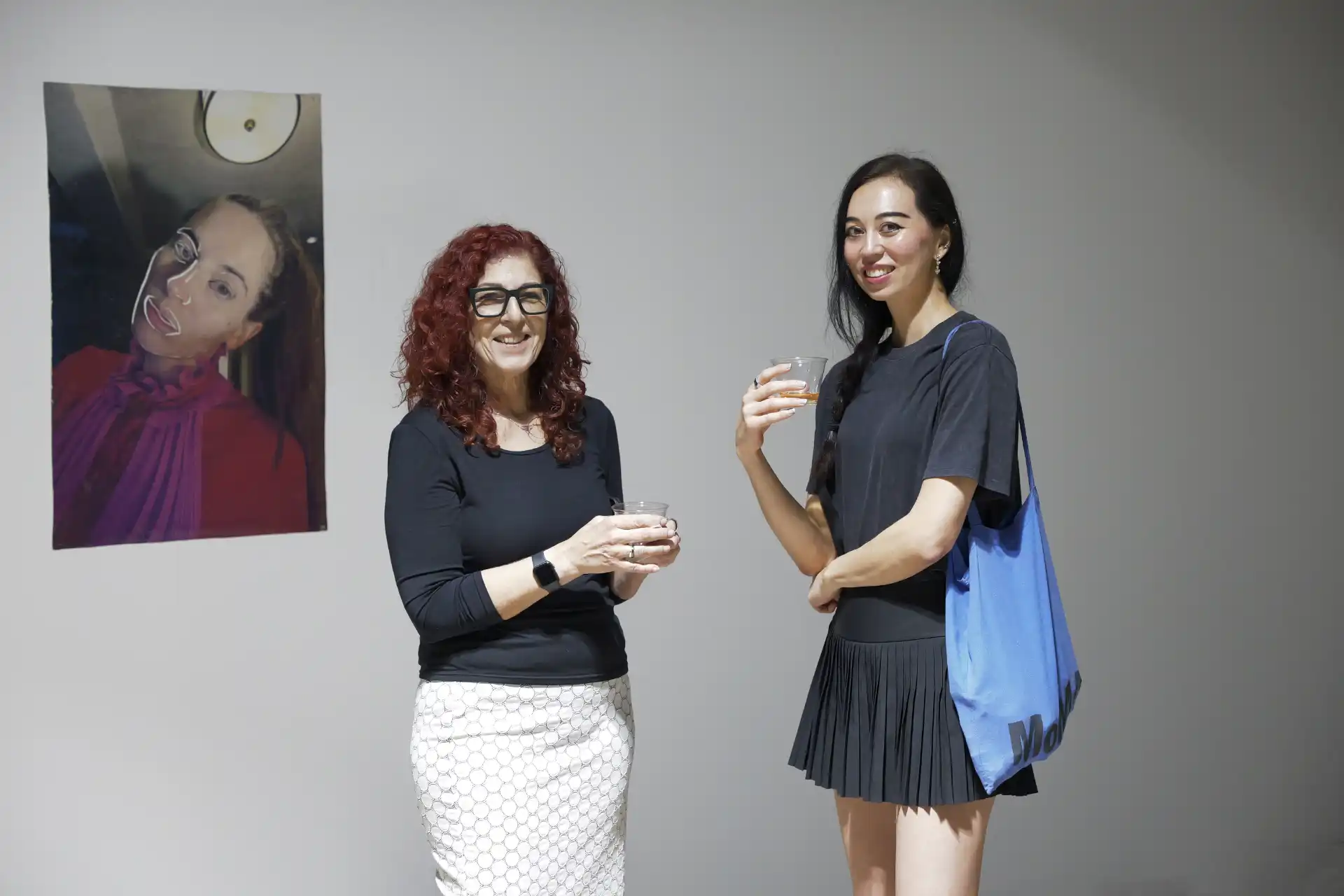 Two women holding drinks stand smiling beside a wall with a portrait of a woman in red and pink.