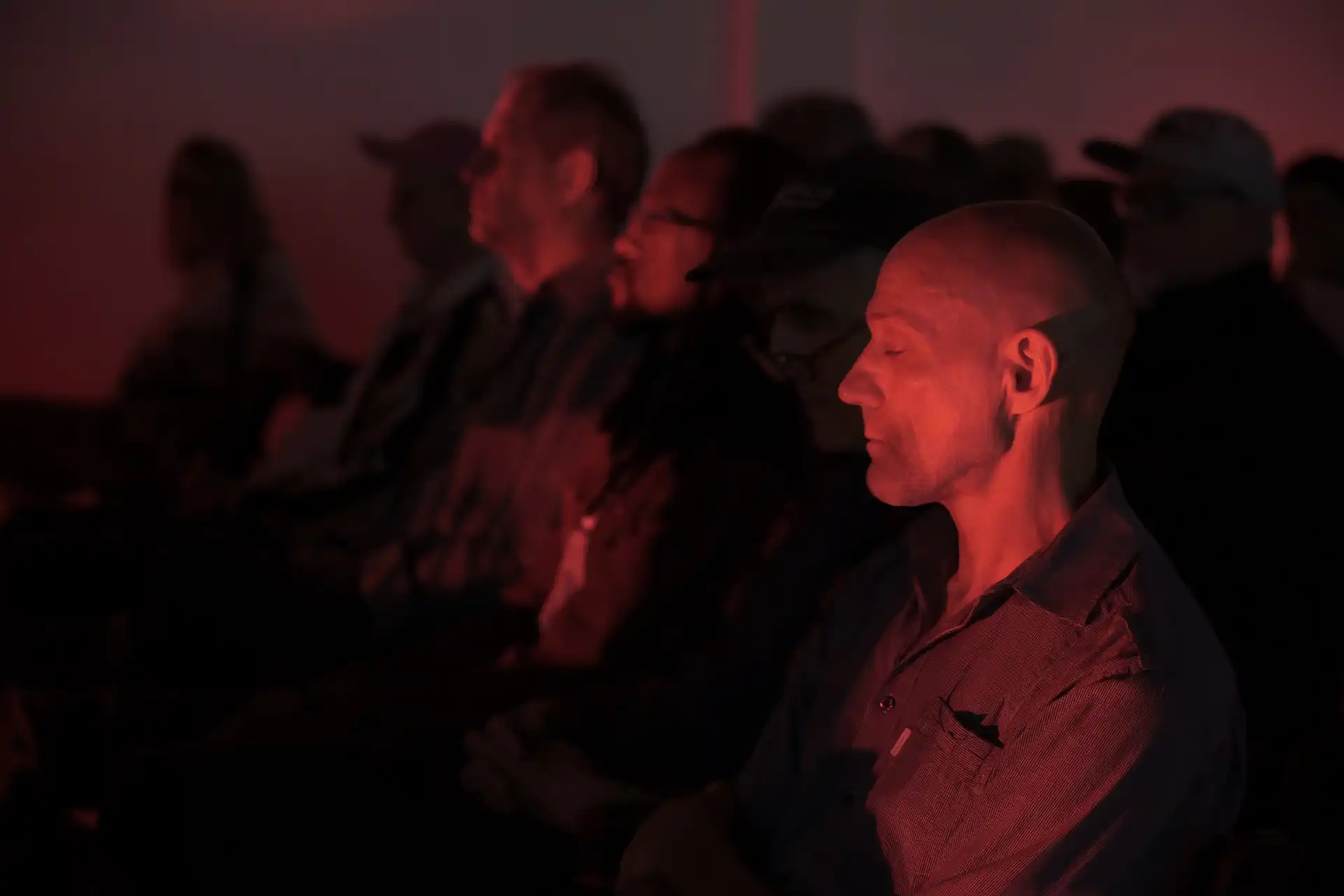 Man with eyes closed sitting in dark room lit by red light among an audience.