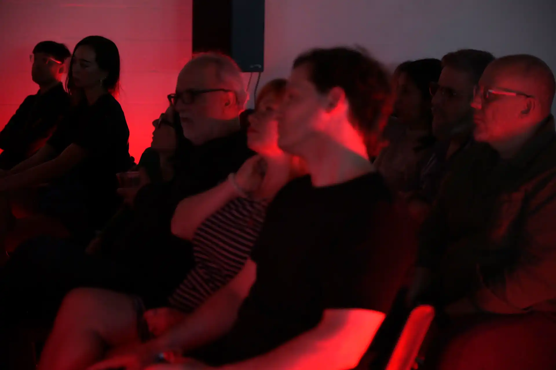 Audience sitting in a dark room lit with red light, focused on an event or performance.