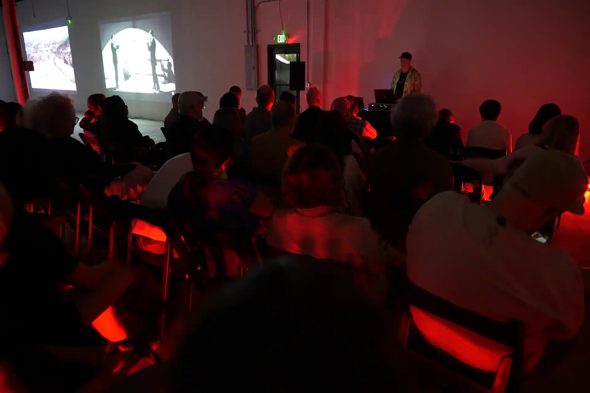 Audience sitting in a dark room watching two black-and-white videos projected on a wall and a presenter at a laptop.