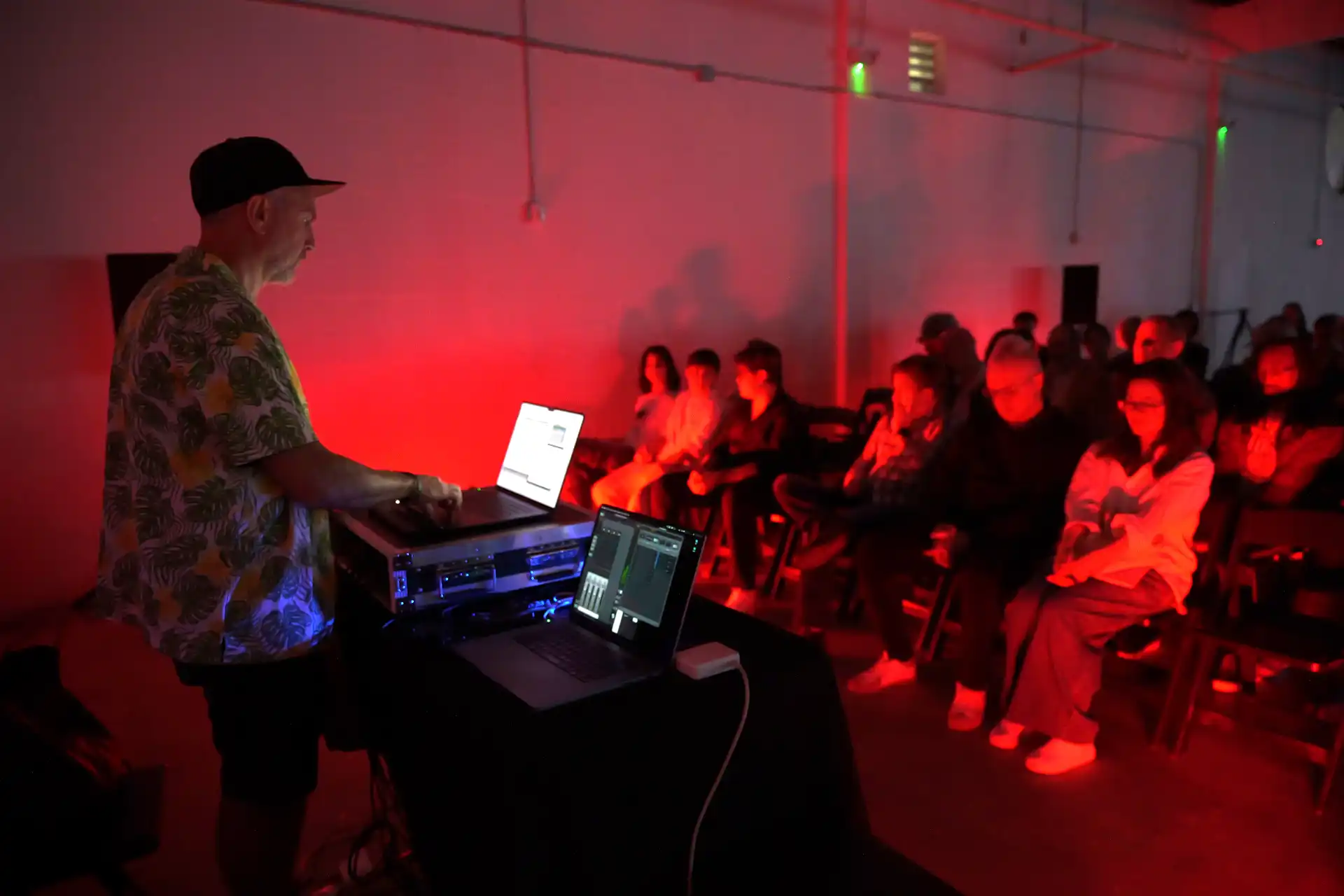 DJ or sound engineer with laptops performing to seated audience bathed in red lighting in a dark room.