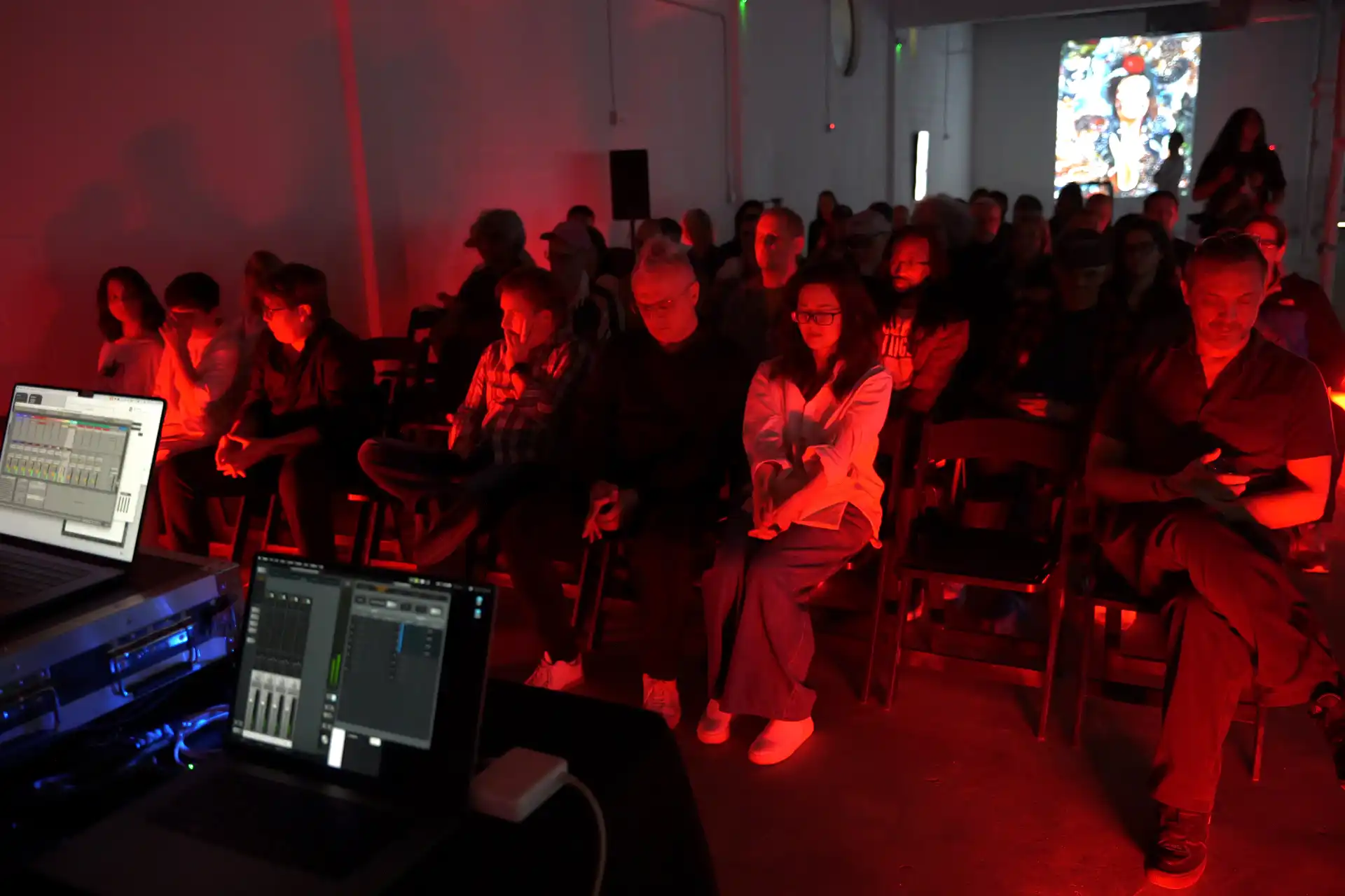 Audience sitting in dark room lit by red light, facing sound mixing equipment with laptops.