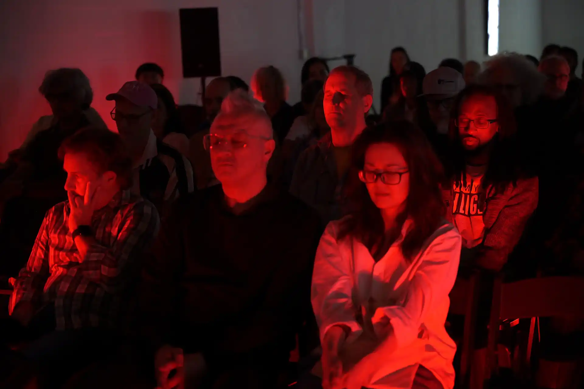 Audience sitting quietly in dim red light at an indoor event, some with eyes closed or deep in thought.