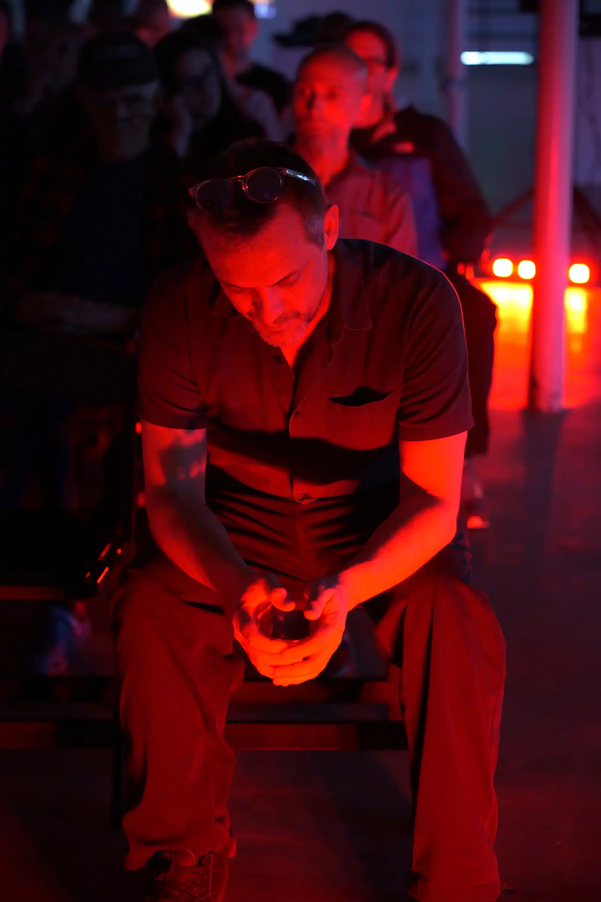 Man sitting with head down, hands clasped, bathed in red light with others blurred behind him.