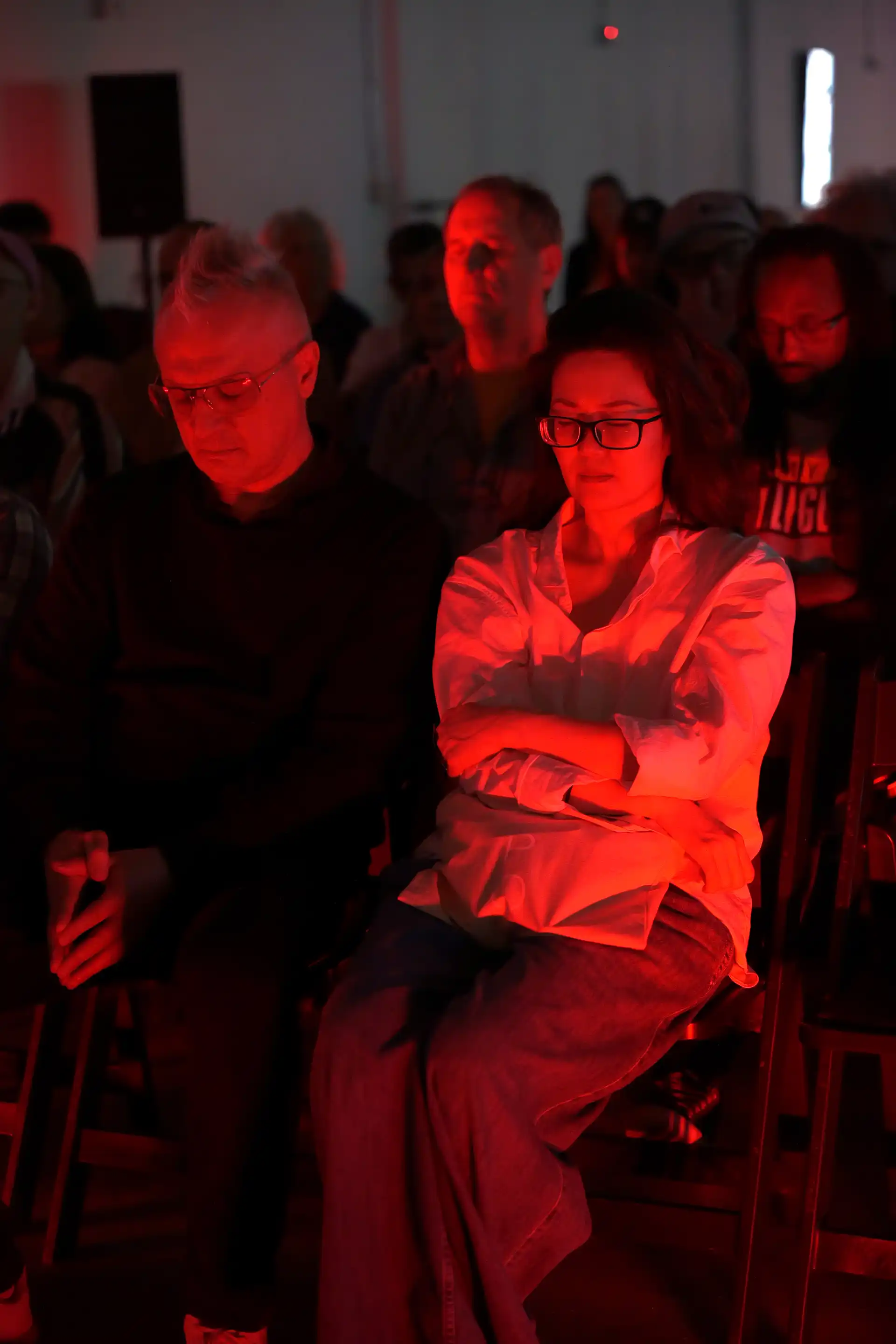 Audience seated with eyes closed, illuminated by red light, appearing calm and focused in a dark room.
