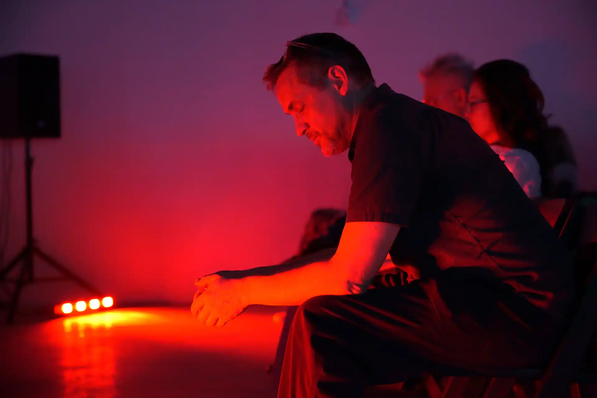 Man sitting with hands clasped, bathed in red light, heads bowed in a dark room with others nearby.