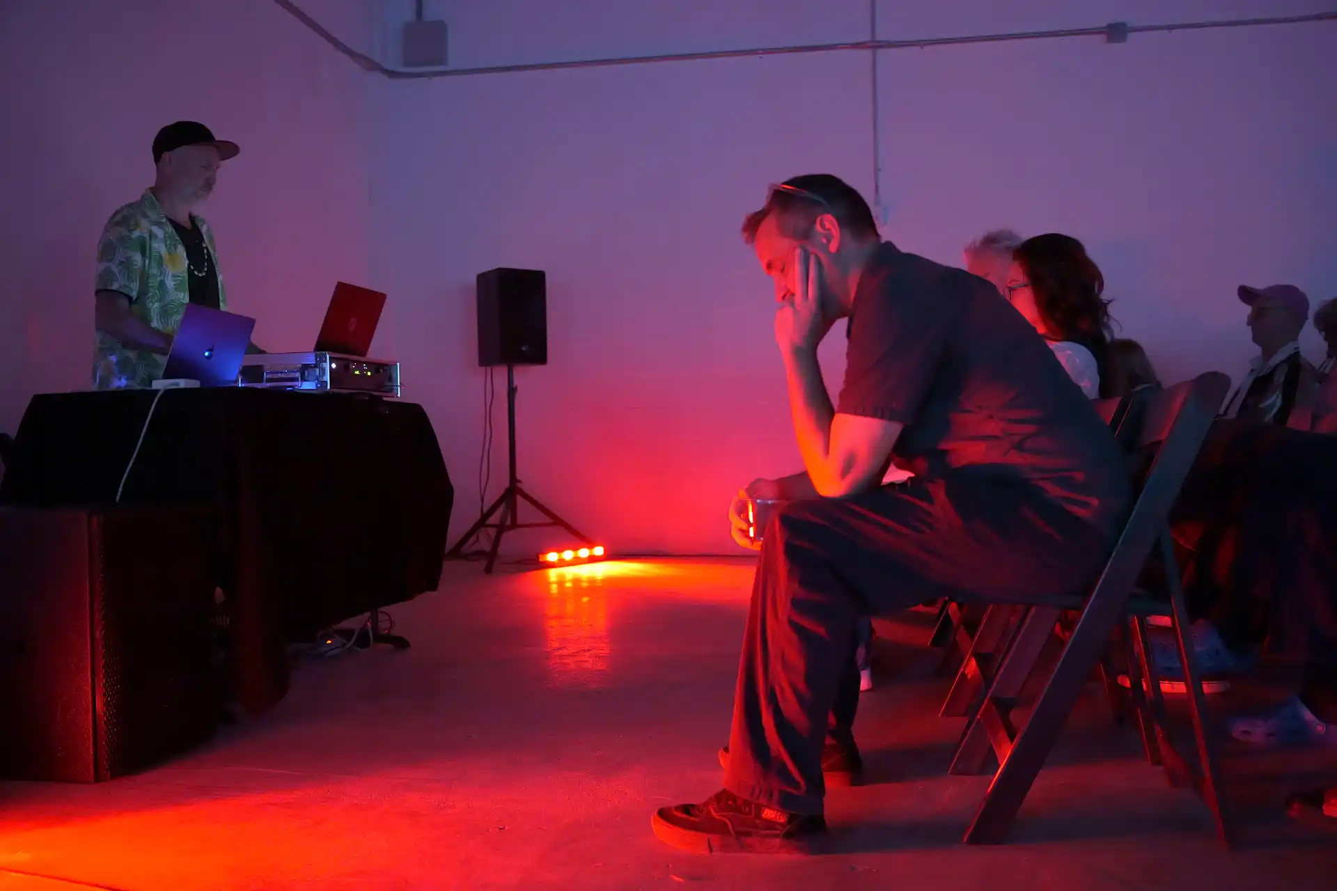 Man in black sits in red-lit room watching DJ with laptops performing at event.
