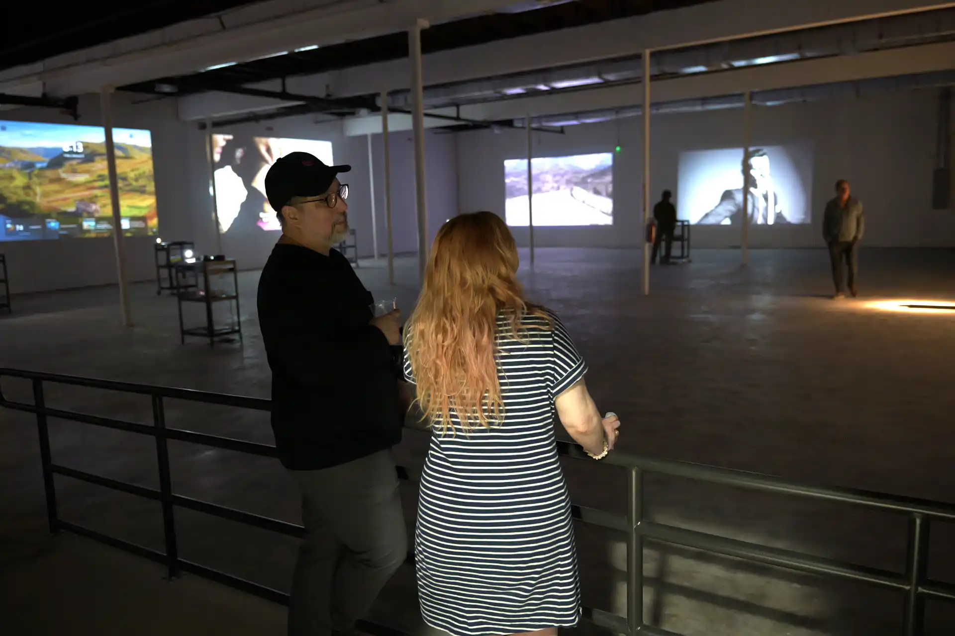 Two people stand by a railing inside a dark gallery with video projections on the walls.