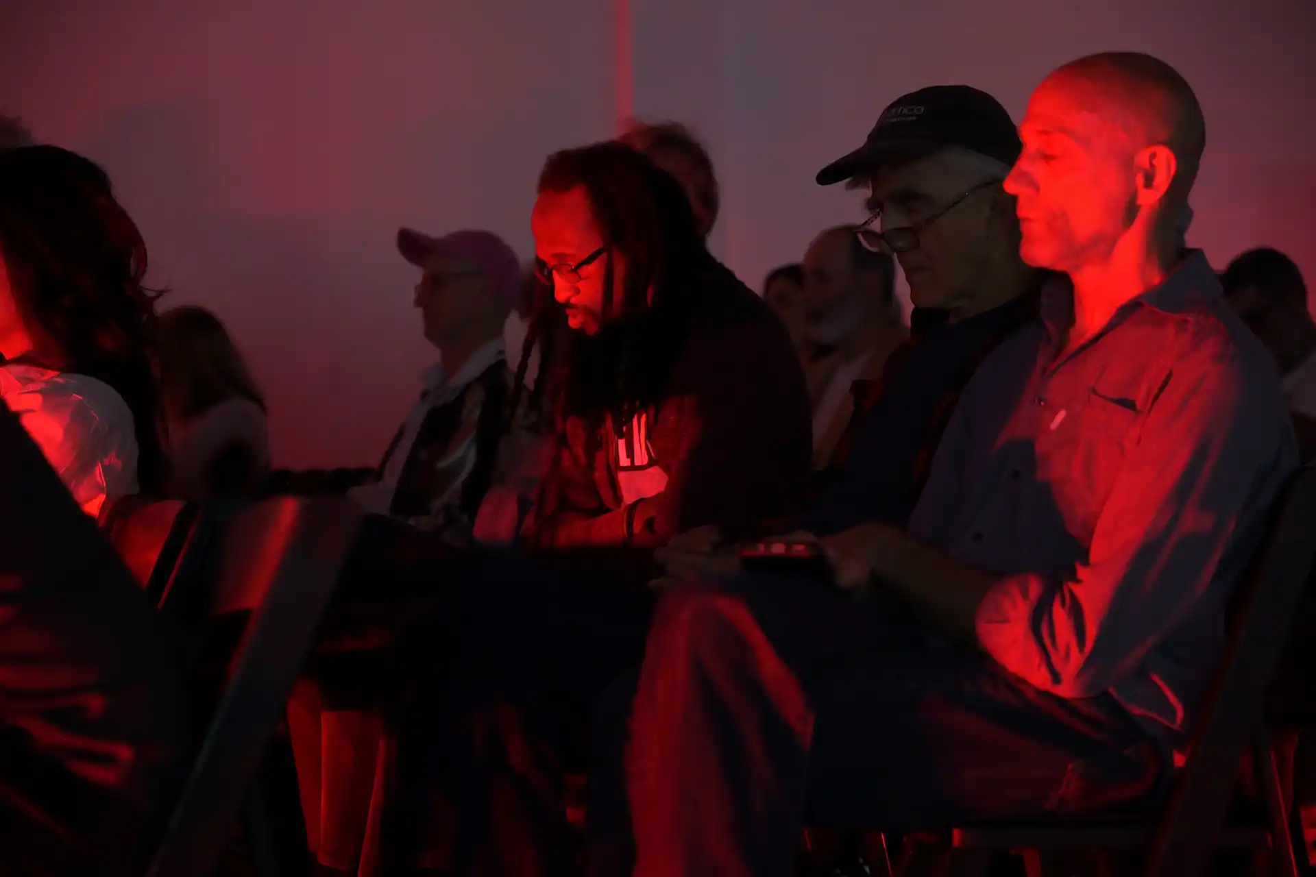 Audience sitting in chairs lit by red light in a dark room, some looking down or at devices.
