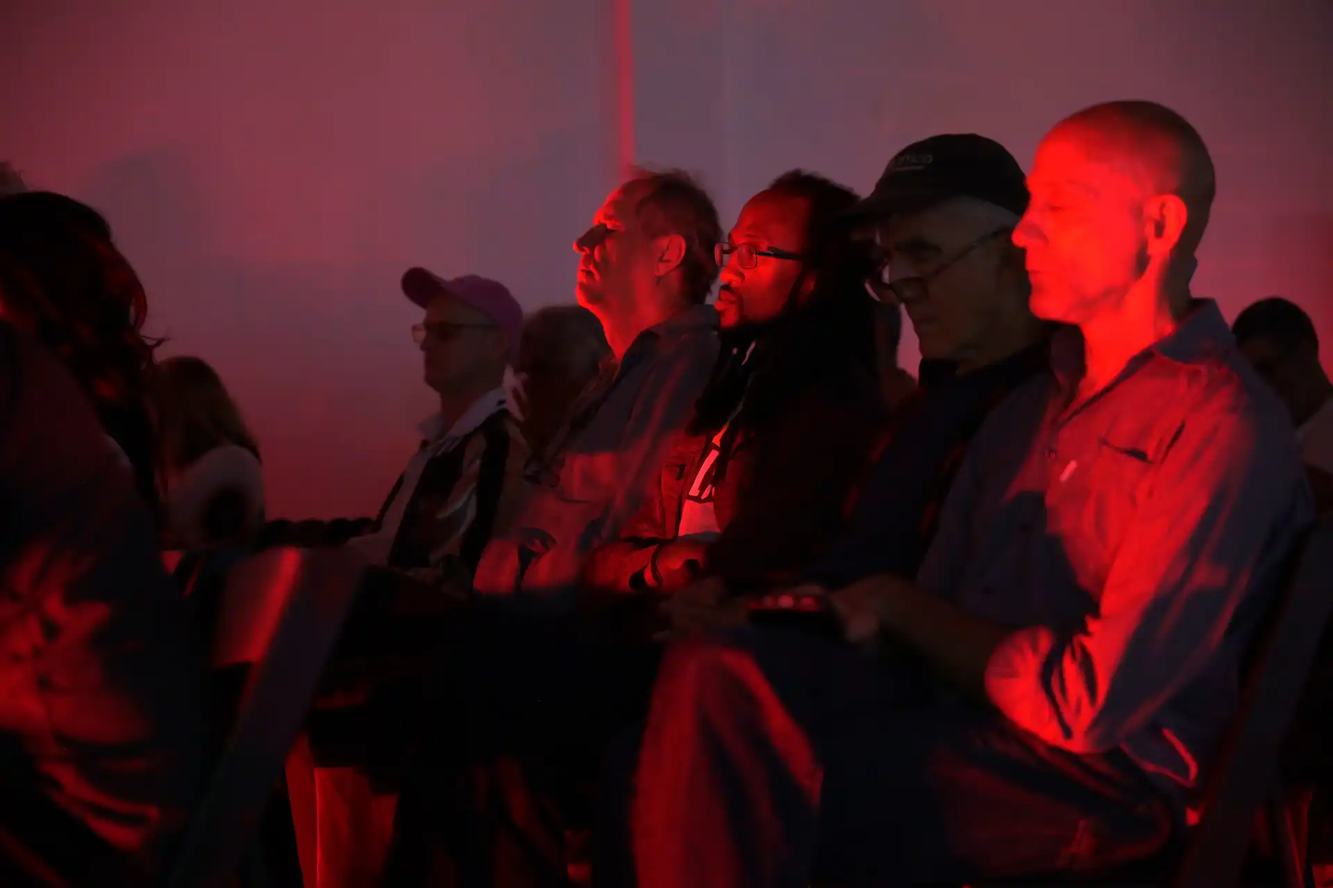 Audience seated in dark room illuminated by red lighting, attentively watching or listening.