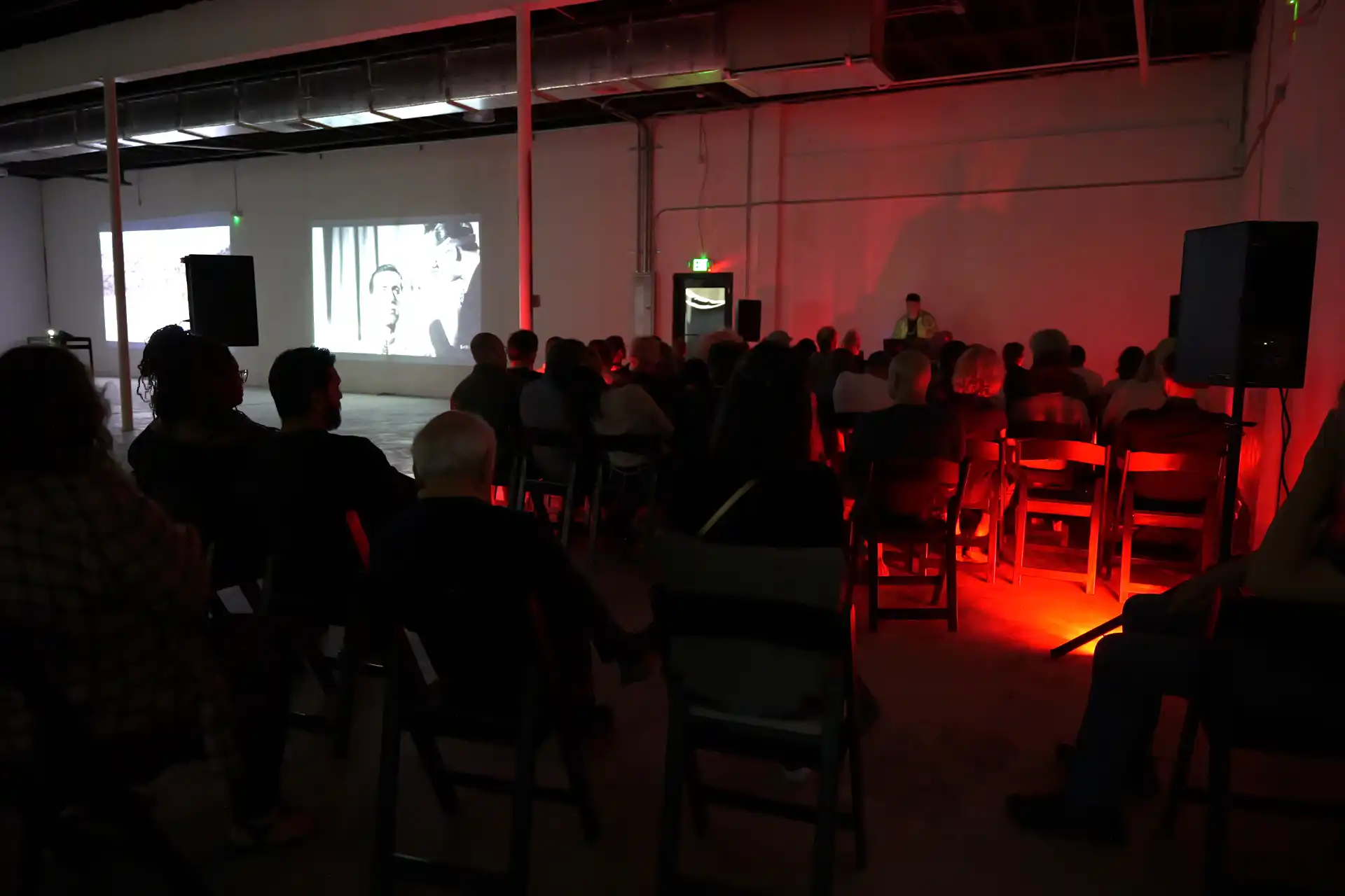 Audience seated in dark room watching black-and-white film projected on a wall with red lighting on right side.