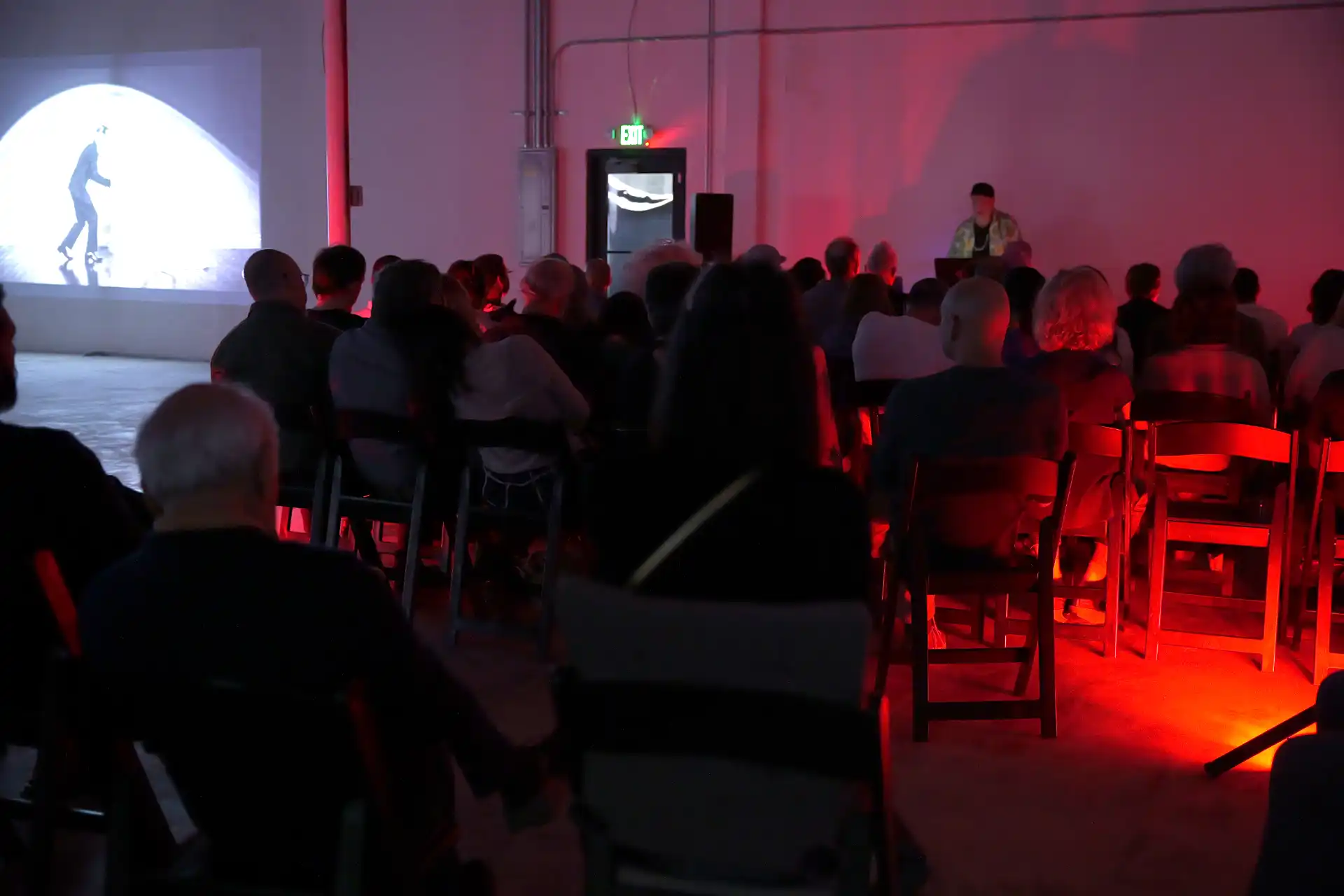 Audience sitting in a dark room watching a DJ and a black-and-white video projected on the wall.