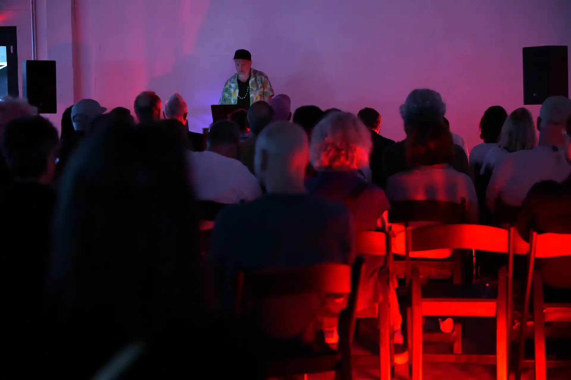 Audience seated and watching a man at a laptop in a dim room with red and purple lighting.