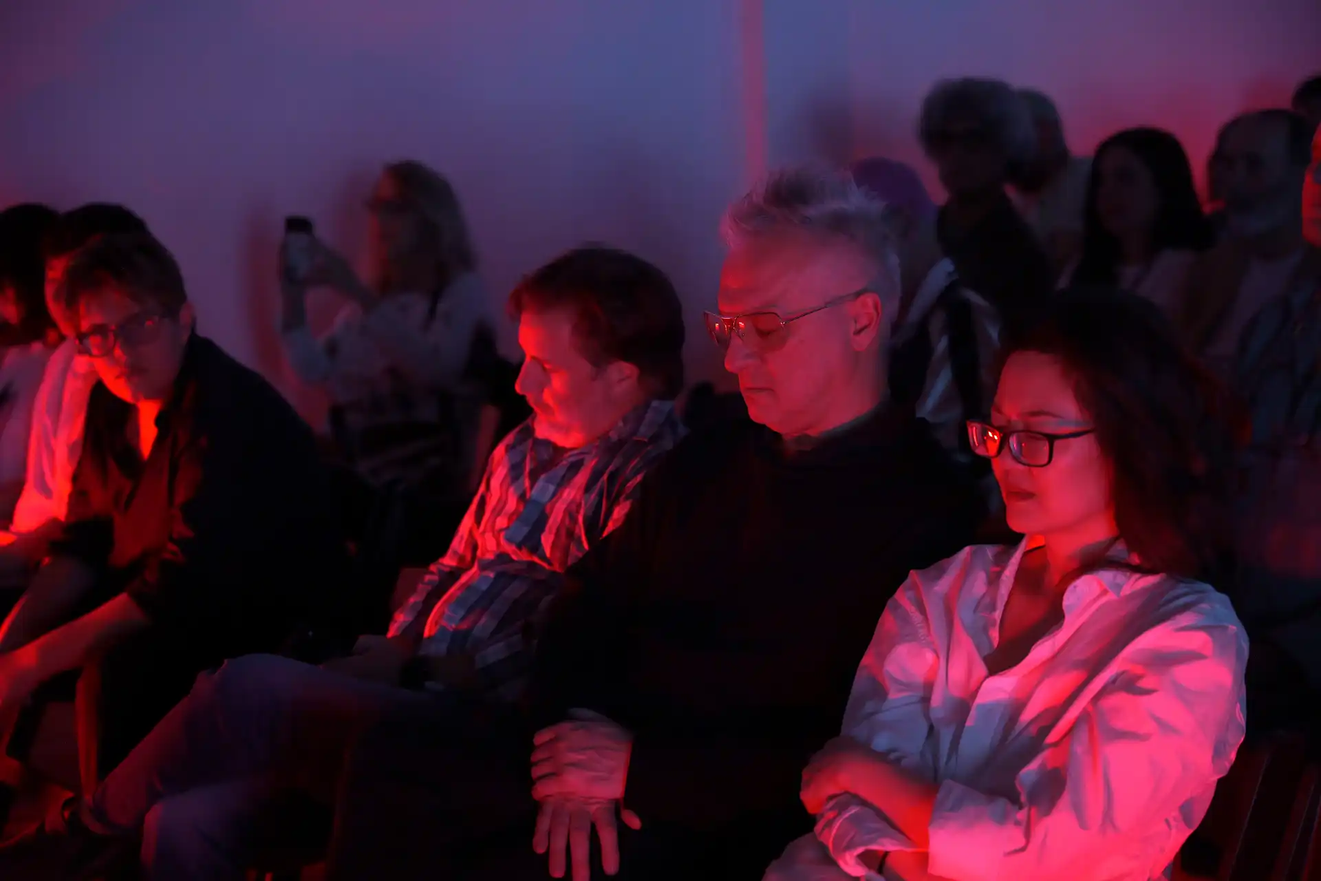 Audience sitting with eyes closed, bathed in red and purple light in a dim room.