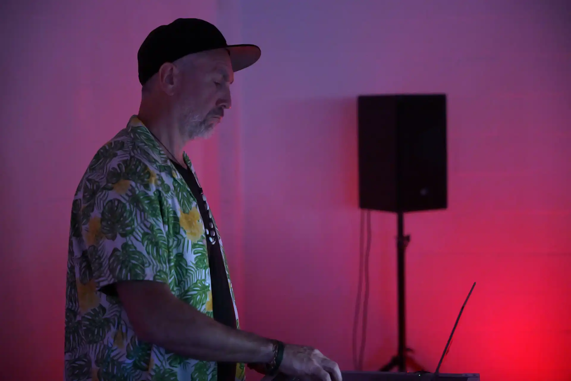 Man in a tropical shirt and cap working on a laptop with purple and red ambient lighting.