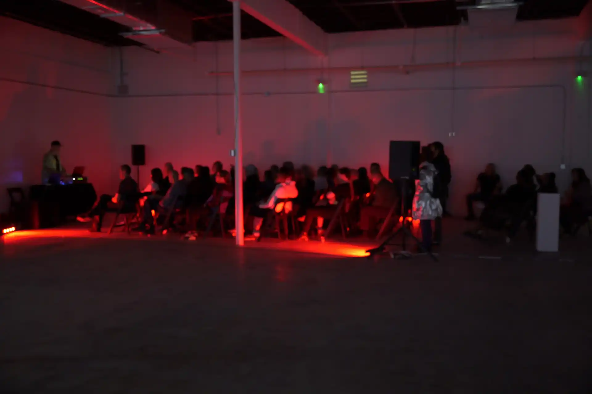 Audience seated in a dimly lit room with red floor lighting and a DJ performing on the left side.