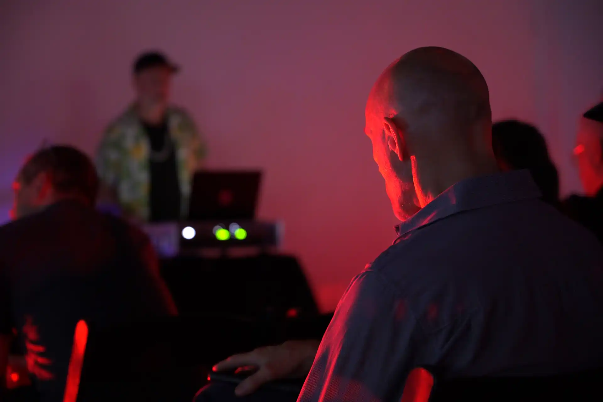 Man sitting in a dim room with red lighting, facing a DJ working on a laptop in the background.