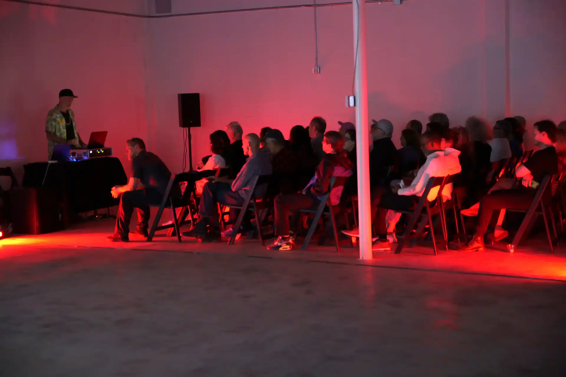 Audience seated in chairs facing a DJ performing with red lighting in a dim room.