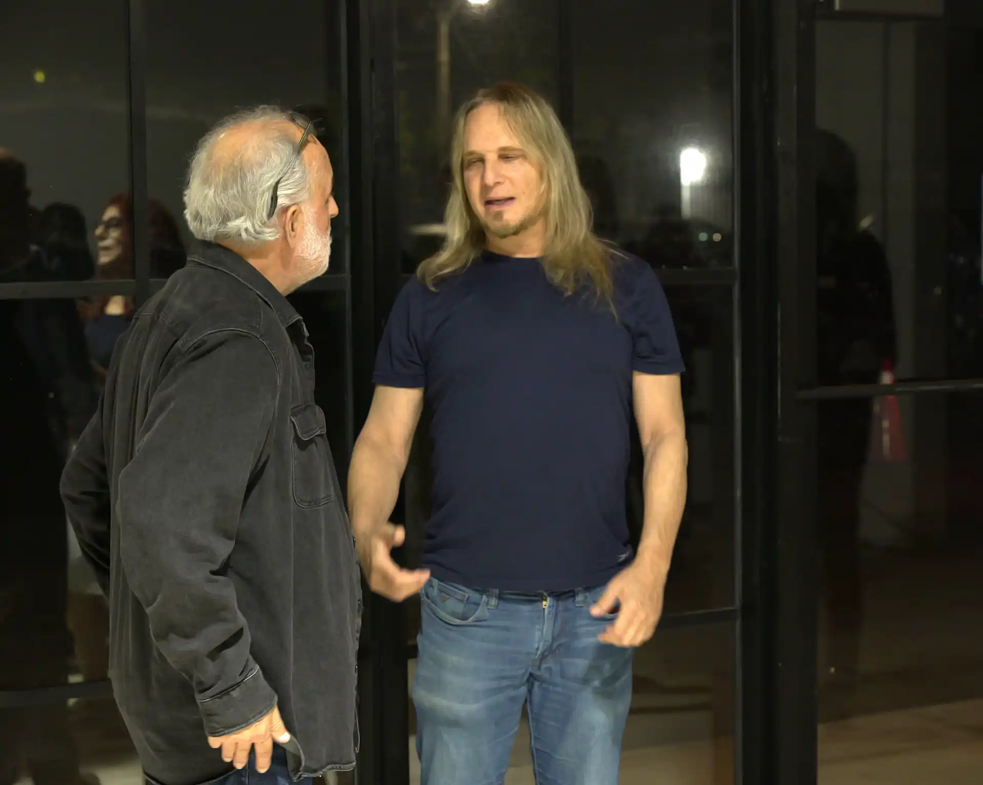 Two men having a conversation indoors at night, one with gray hair and another with long blond hair.