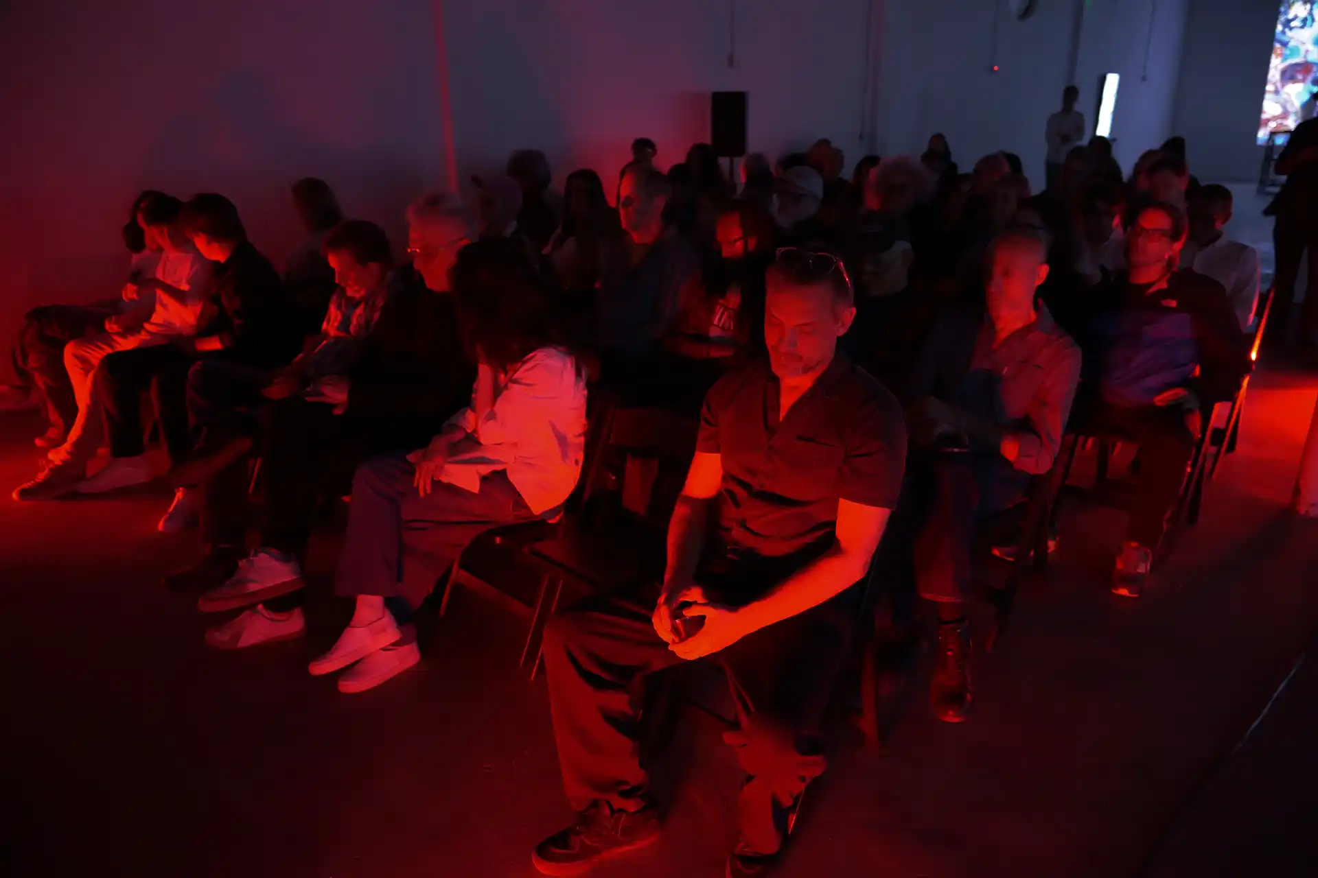 Audience sitting in rows in a dark room illuminated by red light, watching an event or presentation.