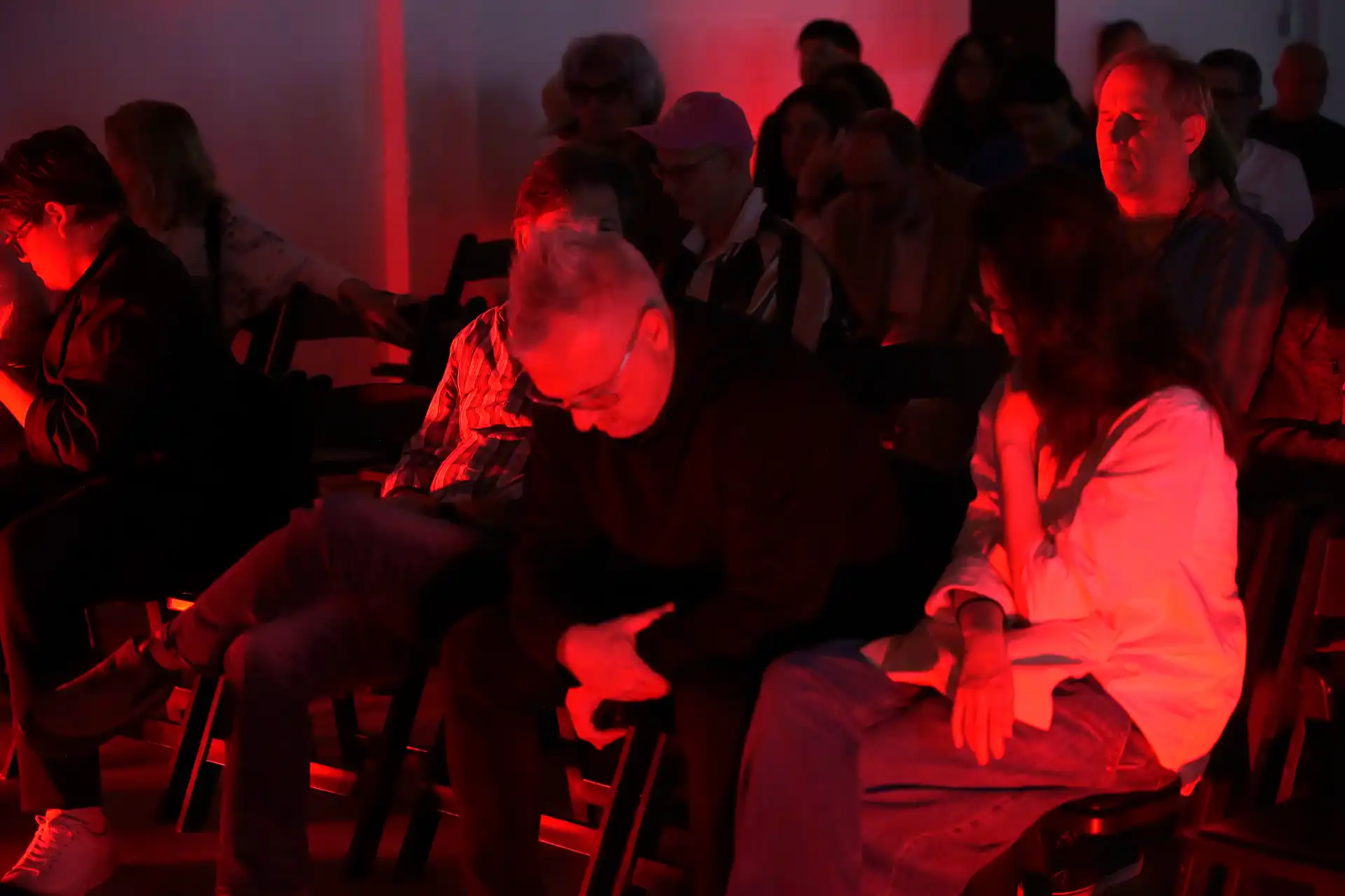 People seated in a dim room bathed in red light, some looking down or with eyes closed.