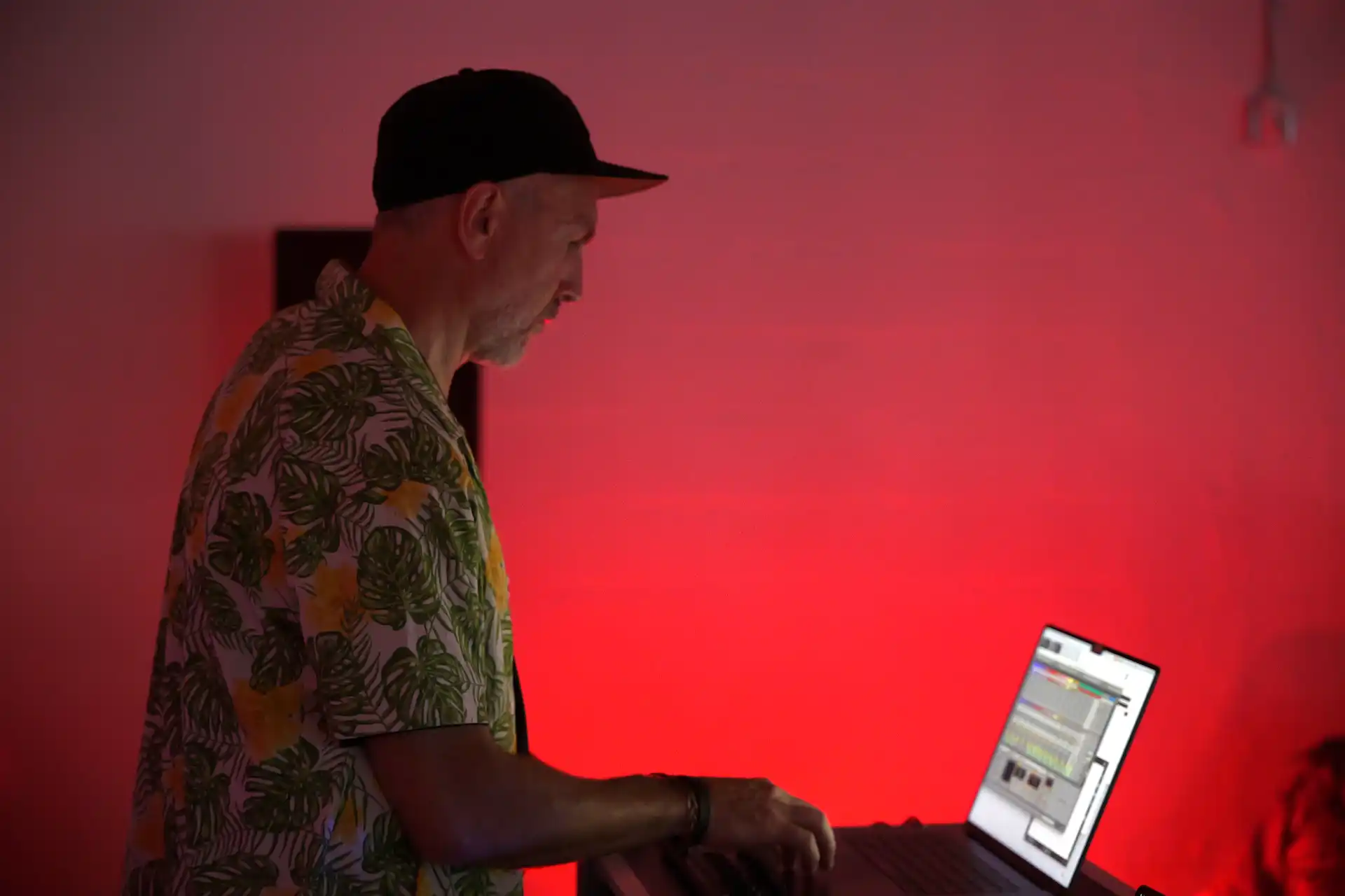 Man in a floral shirt and cap working on a laptop with a red-lit background.