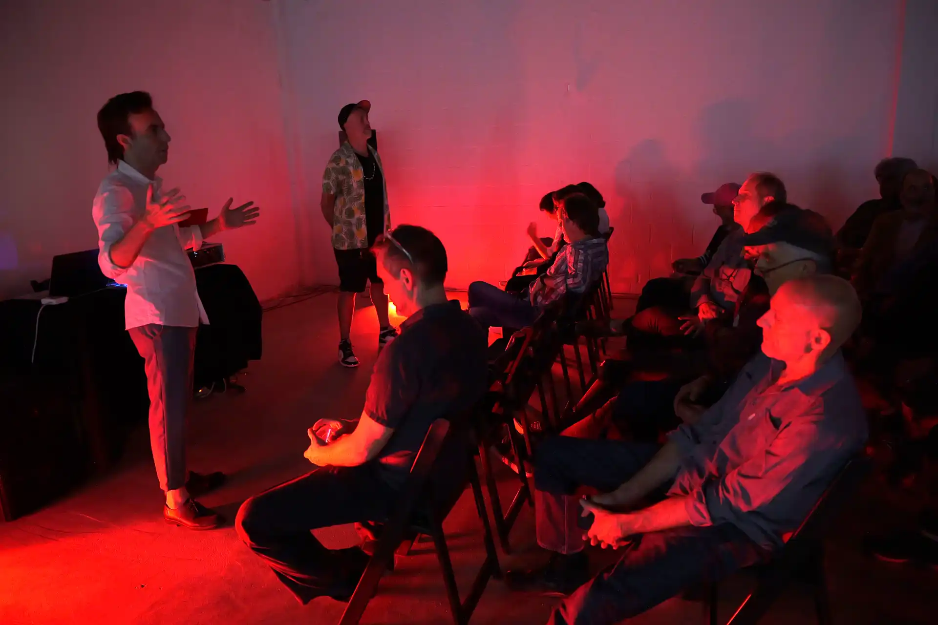 Man speaking to seated audience in dim room with red lighting and casual attire.