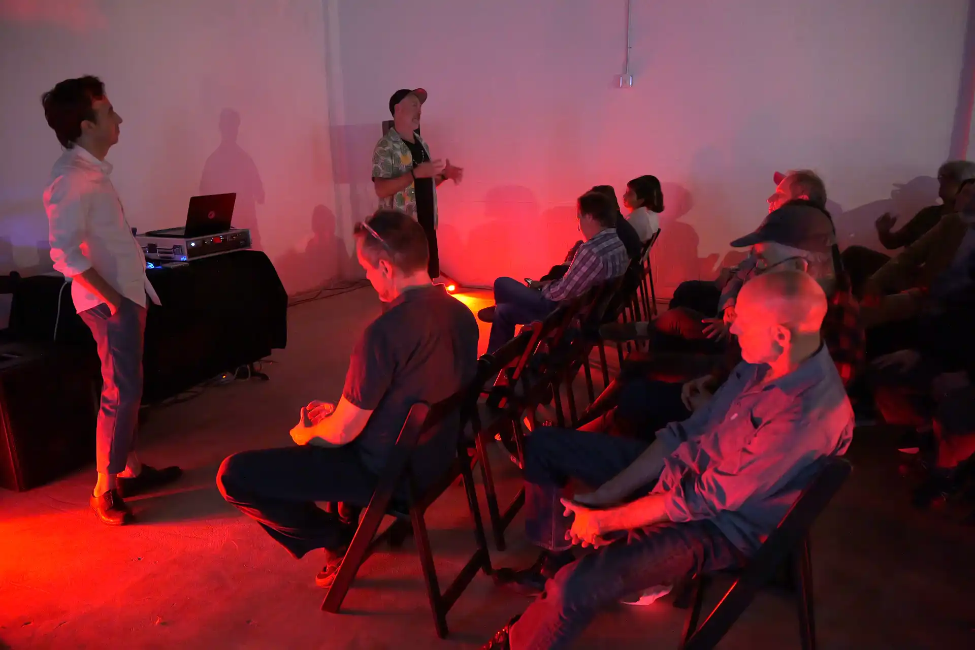 Man in patterned shirt speaking to seated audience in dim room with red lighting.