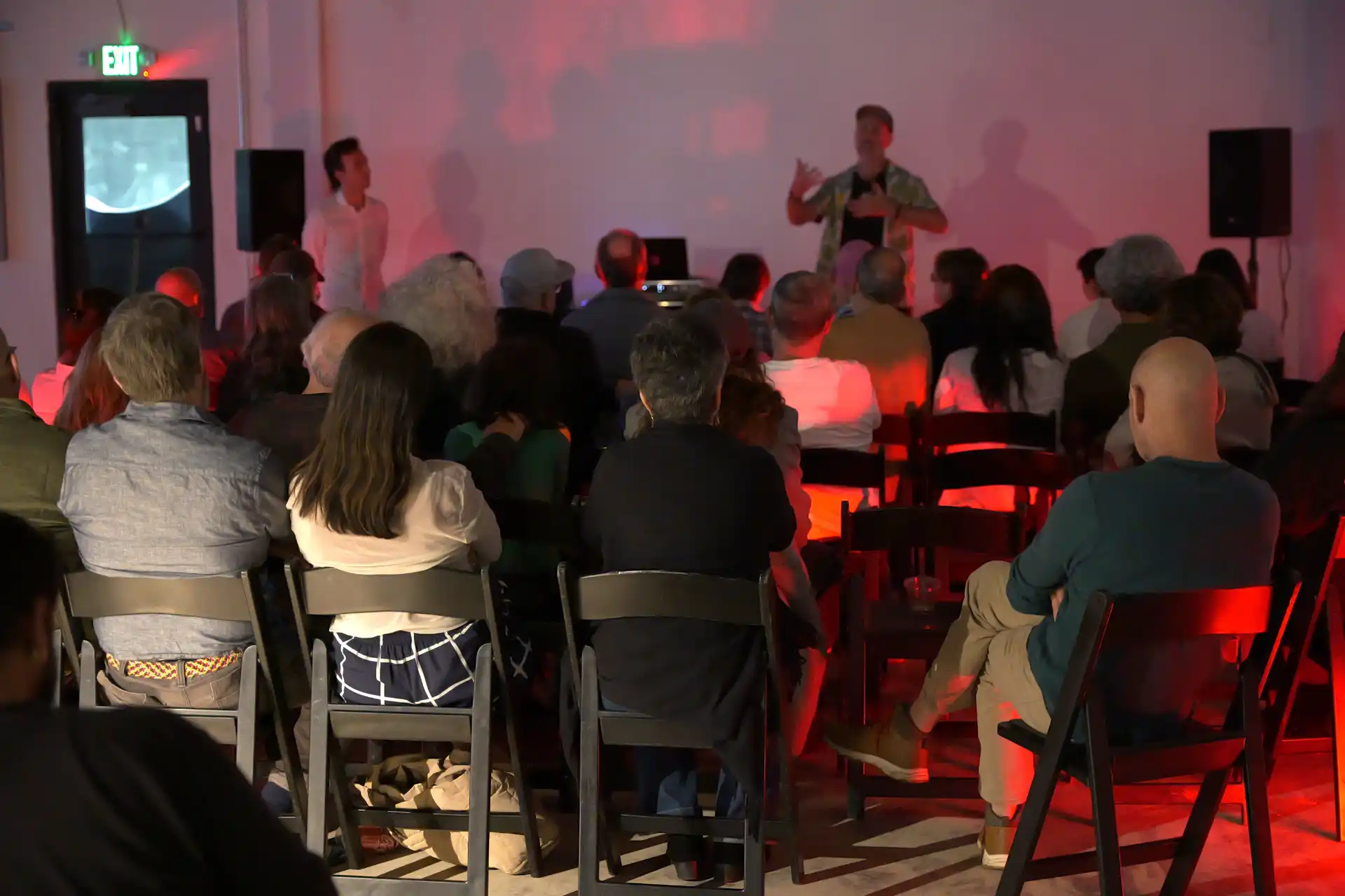 Audience seated in chairs watching a speaker presenting in a dim room with red lighting.
