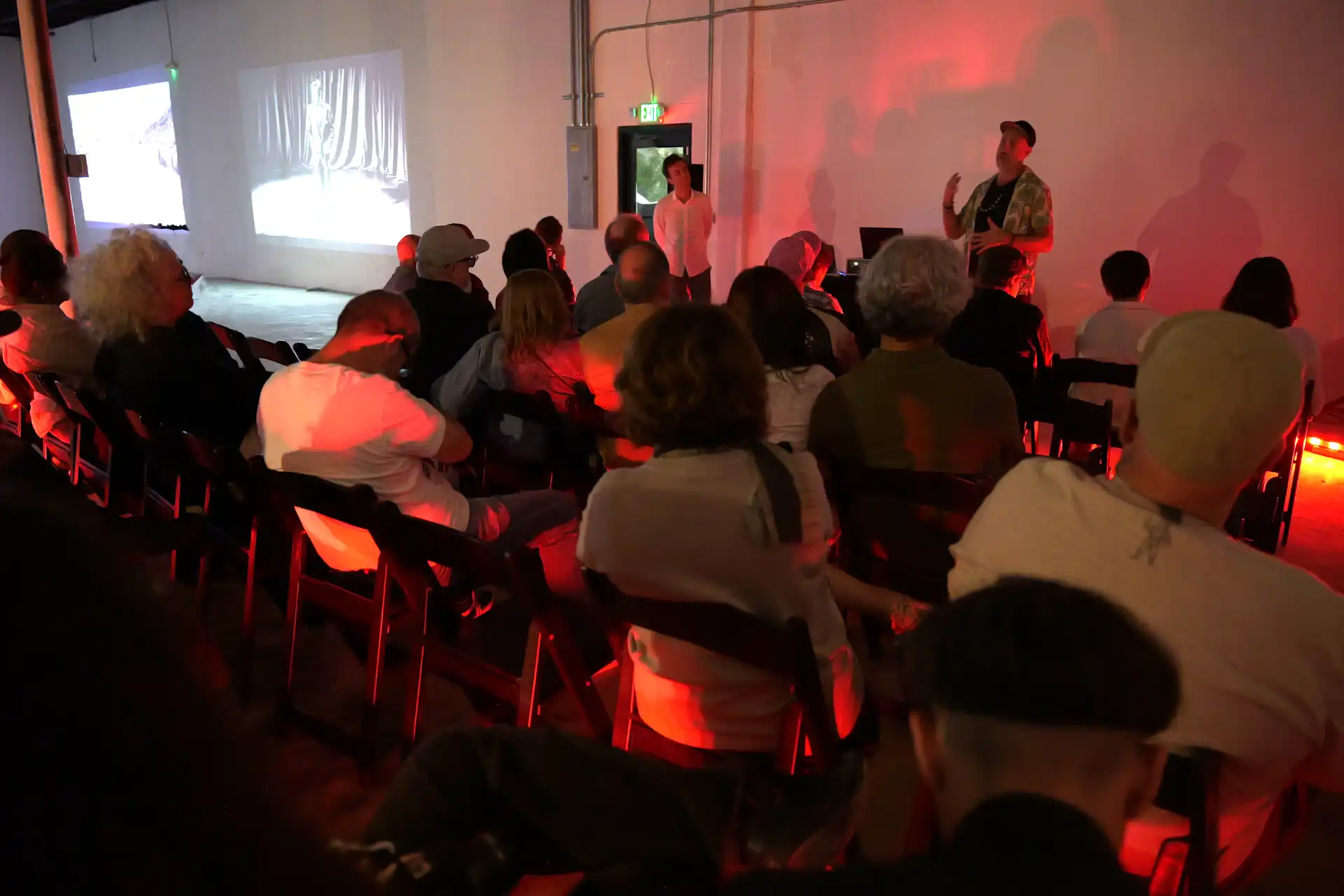 Audience seated watching a speaker in a dim room with red lighting and video projections on the wall.