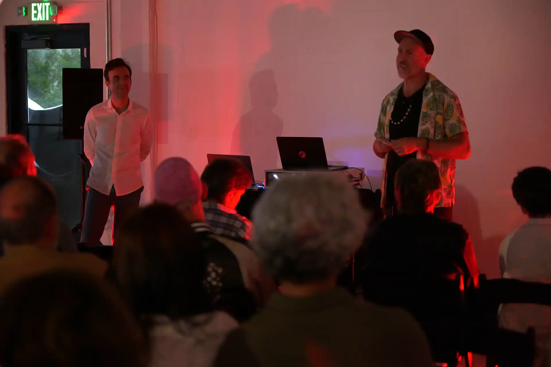 Man in floral shirt speaking to seated audience with red lighting and another man standing by door.