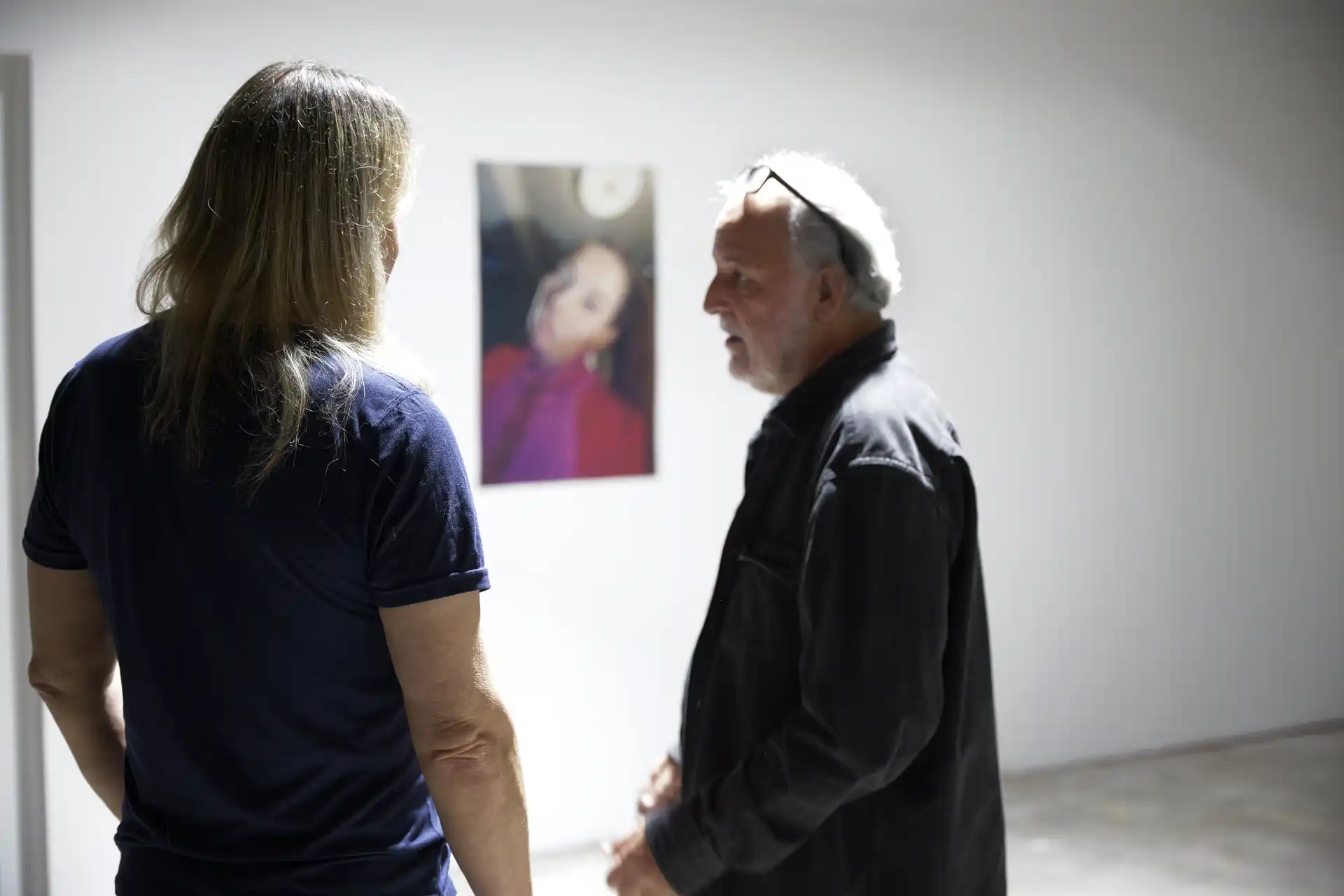 Two men in casual clothes standing indoors, facing a blurred photo on a plain white wall.