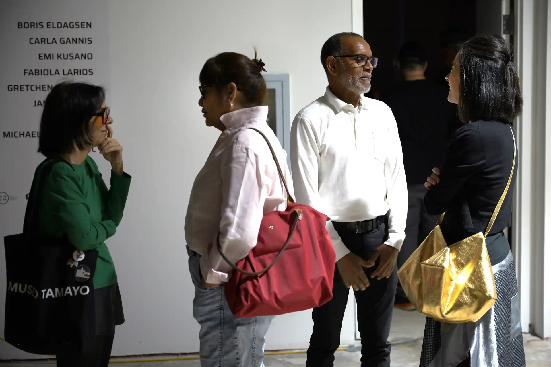 Four people in casual clothes talking in an indoor gallery space with artist names on wall.