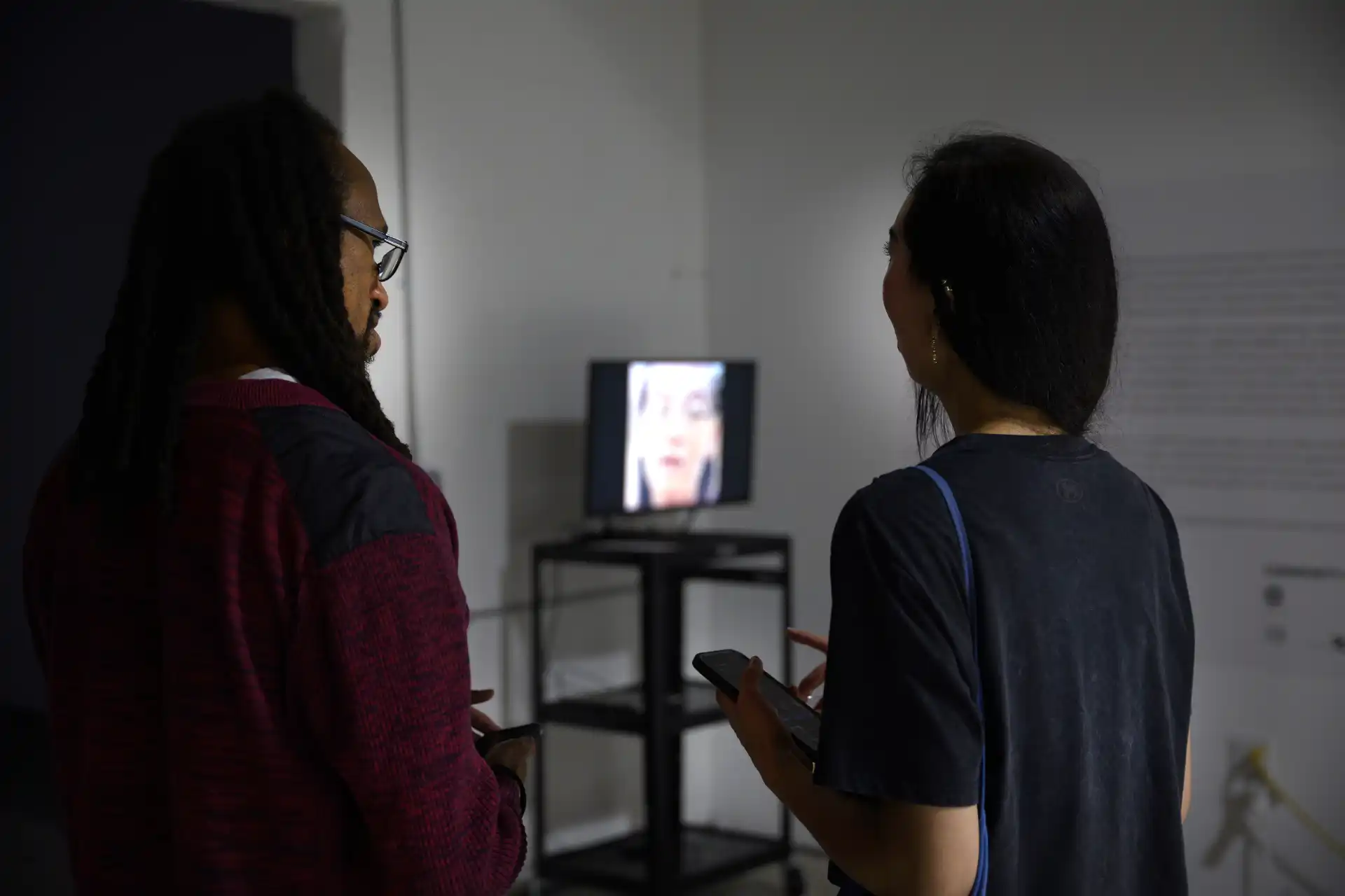 Two people stand in a dim room looking at a blurry face displayed on a screen on a black stand.