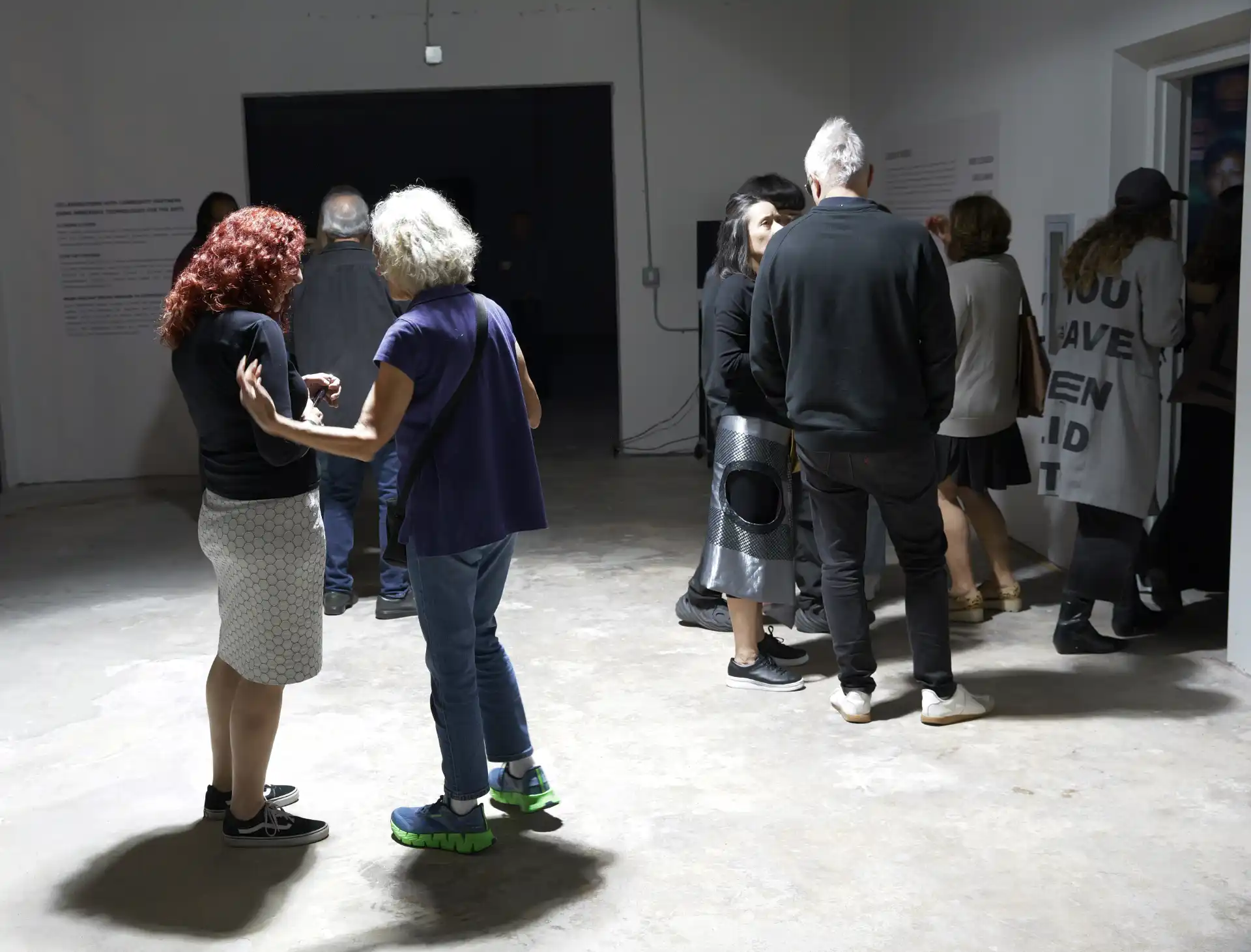 Group of people standing and talking inside a dimly lit gallery space with white walls and concrete floor.